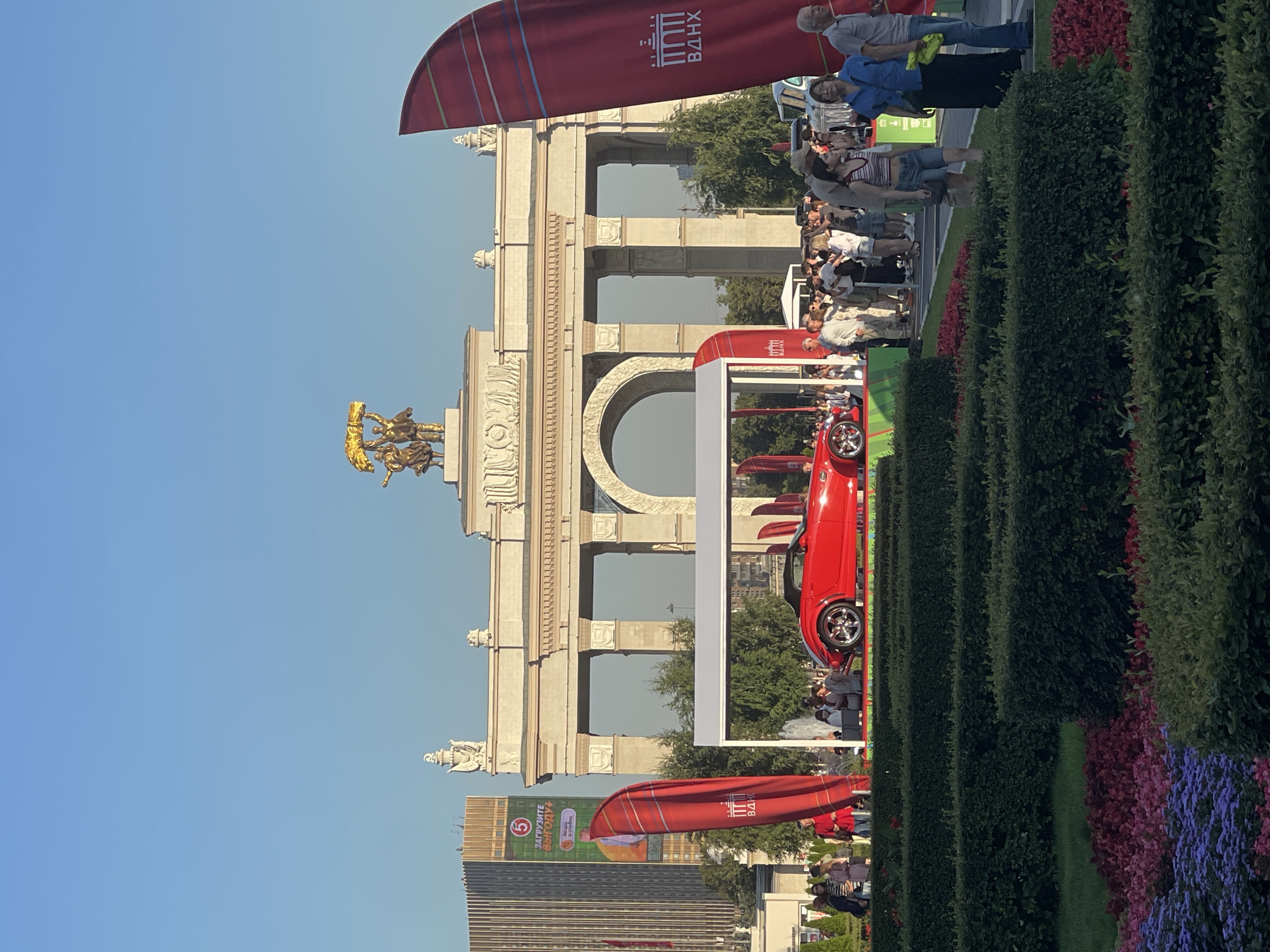 In the city of Unknown, Unknown, an outdoor exhibition features a bright red convertible sports car prominently displayed on a raised platform under a white canopy. The car has a sleek, modern design with chrome accents. Behind the car, a grand archway with multiple columns, topped by a golden sculpture of two figures holding a banner, serves as a prominent architectural landmark. To the left of the archway is a tall, modern building with visible signage. In front of the car, a crowd of people are gathered, observing the vehicle and the surrounding area. Large red banners with Cyrillic lettering are placed on either side of the exhibition space. The foreground is characterized by meticulously manicured green hedges and colorful flowerbeds, with vibrant purple and red blossoms. The sky above is clear and blue, indicating fair weather.