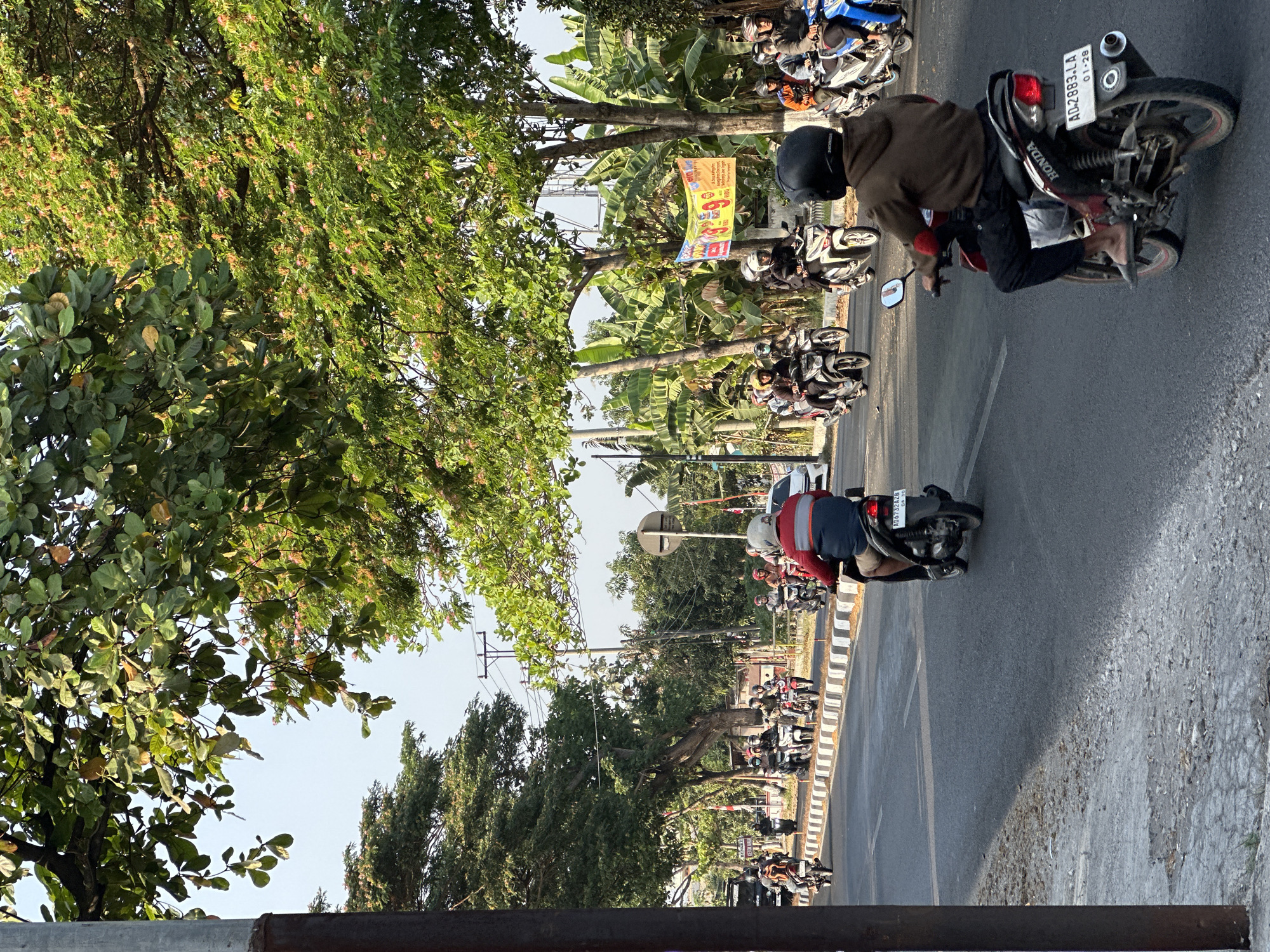 The image depicts a street scene in Bulakrejo, Indonesia, under a bright sky, with a line of trees casting shadows across the asphalt road. A group of people on motorcycles are traveling in the same direction. The foreground features a person riding a Honda motorcycle with the license plate AD-2883-LA. They are wearing a dark helmet and a brown hooded sweatshirt. Further down the road, other motorcyclists are visible, some wearing helmets and jackets. The roadside is lined with a white and black striped curb, with vegetation and trees beyond. A banner is hanging from a pole on the right side of the road. The overall impression is of a sunny day with moderate traffic.