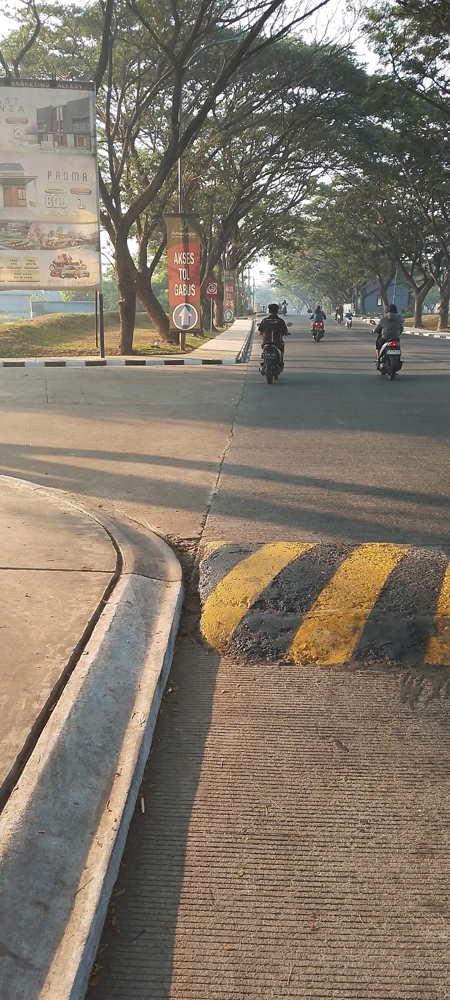 This is a street-level, eye-level, vertical shot taken at a slight upward angle on a paved road in Satriamekar, Indonesia. The image depicts a sunny day with long shadows cast across the asphalt, indicating either early morning or late afternoon. A concrete curb with a drainage channel is visible in the foreground to the left, and a speed bump with black and yellow diagonal stripes is in the middle ground.
Several motorcycles are on the road, all traveling in the same direction away from the viewer. The closest motorcycle on the right has a rider wearing a grey jacket and helmet. Further down the road, another motorcycle has a rider in a black and red jacket and helmet. Several other motorcycles are visible in the distance.
Along the left side of the road, there is a sidewalk bordered by a white and black striped curb. Behind the sidewalk are trees with dense green foliage, casting dappled shadows. Several signs are posted along the sidewalk. One large banner, partially visible on the far left, appears to be advertising real estate. A red and white sign with Indonesian text and a circular arrow pointing upwards is also visible, along with a 40 km/h speed limit sign and another identical banner sign further down the road. The overall environment suggests a residential or developing area with tree-lined streets.