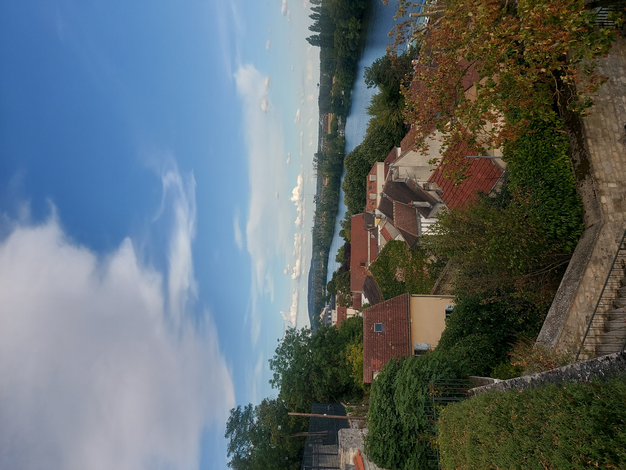 This is a wide-angle, high-angle shot of a riverside town in Conflans-Sainte-Honorine, France, on a clear day. The foreground is dominated by a stone wall and lush green hedges. Behind the hedges, a cluster of red-tiled roofs of houses is visible. A winding path with stone steps and a metal railing descends from the foreground.
Beyond the houses, a wide, blue river, likely the Seine, stretches into the distance. The riverbanks are lined with dense green trees, and further in the background, more trees and buildings can be seen, suggesting a developed urban or semi-urban area. The sky is a vibrant blue, punctuated by scattered white clouds. The overall atmosphere is serene and picturesque, capturing a typical French riverside town scene. No people or animals are visible in the image. The lighting indicates it is daytime, with the sun casting soft shadows.