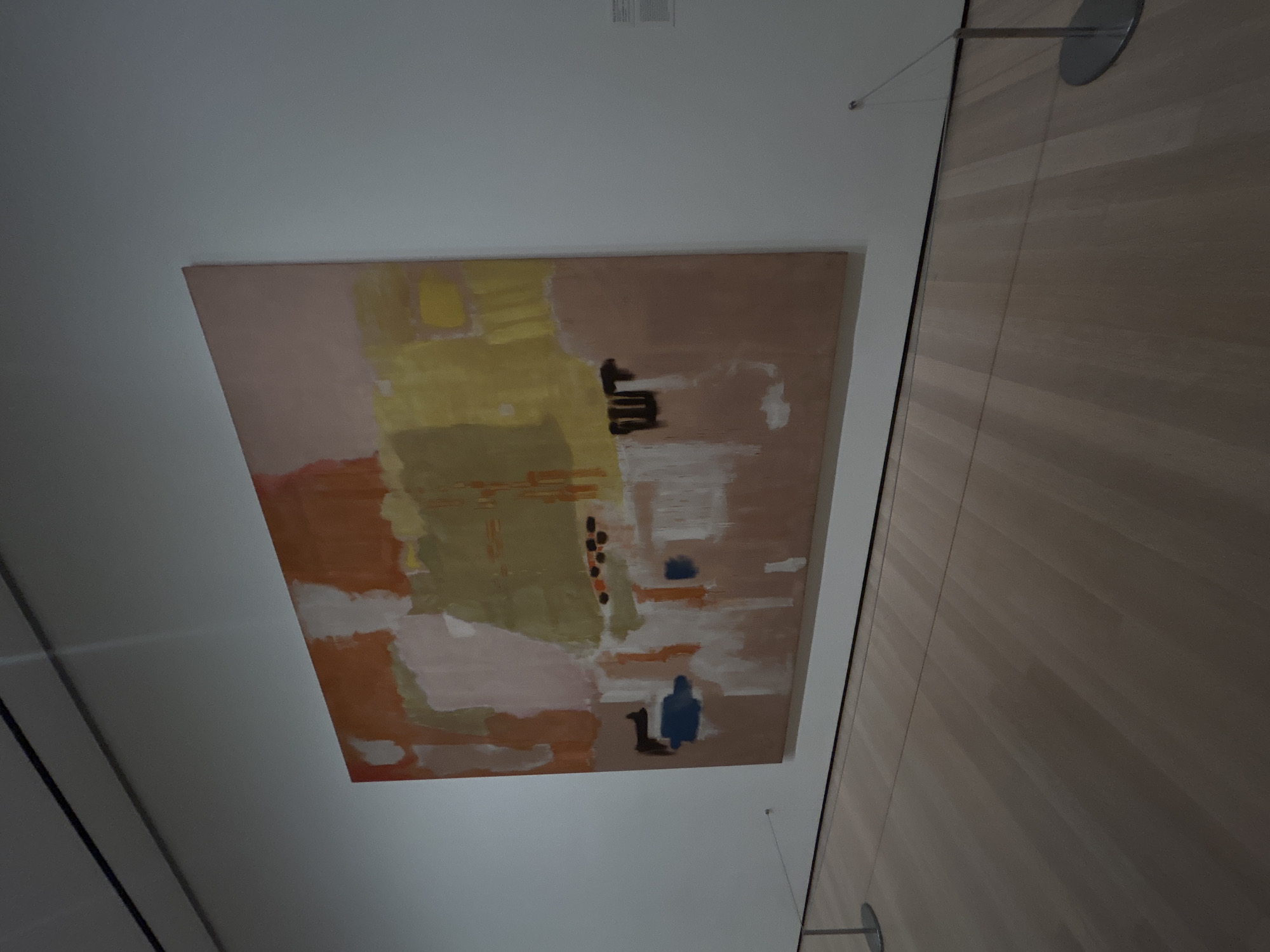 This media file captures a large, rectangular abstract painting displayed on a plain white wall within an indoor setting, likely an art gallery or museum in New York, United States of America. The painting features broad, expressive brushstrokes and large color fields. Dominant colors include muted yellows, olive greens, various shades of brown and dusty rose, and splashes of orange-red and white.
Within the abstract composition, several distinct elements are visible. Towards the upper mid-right, there is a prominent vertical black abstract shape, possibly a stylized symbol or series of strokes. Below and slightly to the left of this black shape, a cluster of small, dark brown or black dots is present. In the bottom right quadrant of the painting, a significant blue, blob-like figure is depicted alongside a smaller, dark, stylized silhouette that resembles a duck or a quadrupedal animal. Another smaller blue shape is visible above the main blue figure.
The perspective of the shot is slightly angled, looking upwards at the artwork. To the right of the painting, a section of a light brown, vertically paneled wall or door is visible, extending from the top to the bottom of the frame. Thin wires, likely part of the display system, can be seen extending from the upper right, attached to a silver, circular fixture on the wall. A small, blurry white rectangle containing illegible dark text, presumably an exhibition label or information plaque, is visible on the white wall above the painting. The lighting is uniform, indicative of artificial indoor illumination. The scene captures the static display of contemporary art.