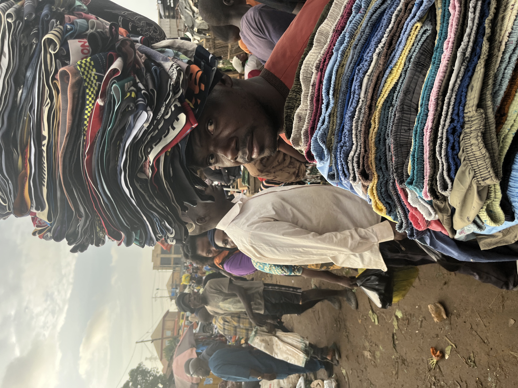 A man with a stack of folded clothes on his head is the central subject of this image, taken in the city of Jos, Nigeria. He is looking directly at the camera with a serious expression. He is wearing an orange shirt. In front of him, he is carrying another stack of folded clothes, appearing to be shorts or pants, which are brightly colored with blues, yellows, pinks, and grays.
The background reveals a busy market scene. Several other people are visible, some walking and others standing. To the left, a man in a blue garment is walking with a white bag. Another man in a grey t-shirt and dark shorts is also walking in the mid-ground. In the distance, there are buildings and market stalls, with umbrellas and awnings suggesting a bustling commercial area. The ground is dirt and appears somewhat uneven, with scattered debris. The sky above is overcast with hints of sunlight breaking through. The image captures a dynamic moment in a lively Nigerian marketplace.