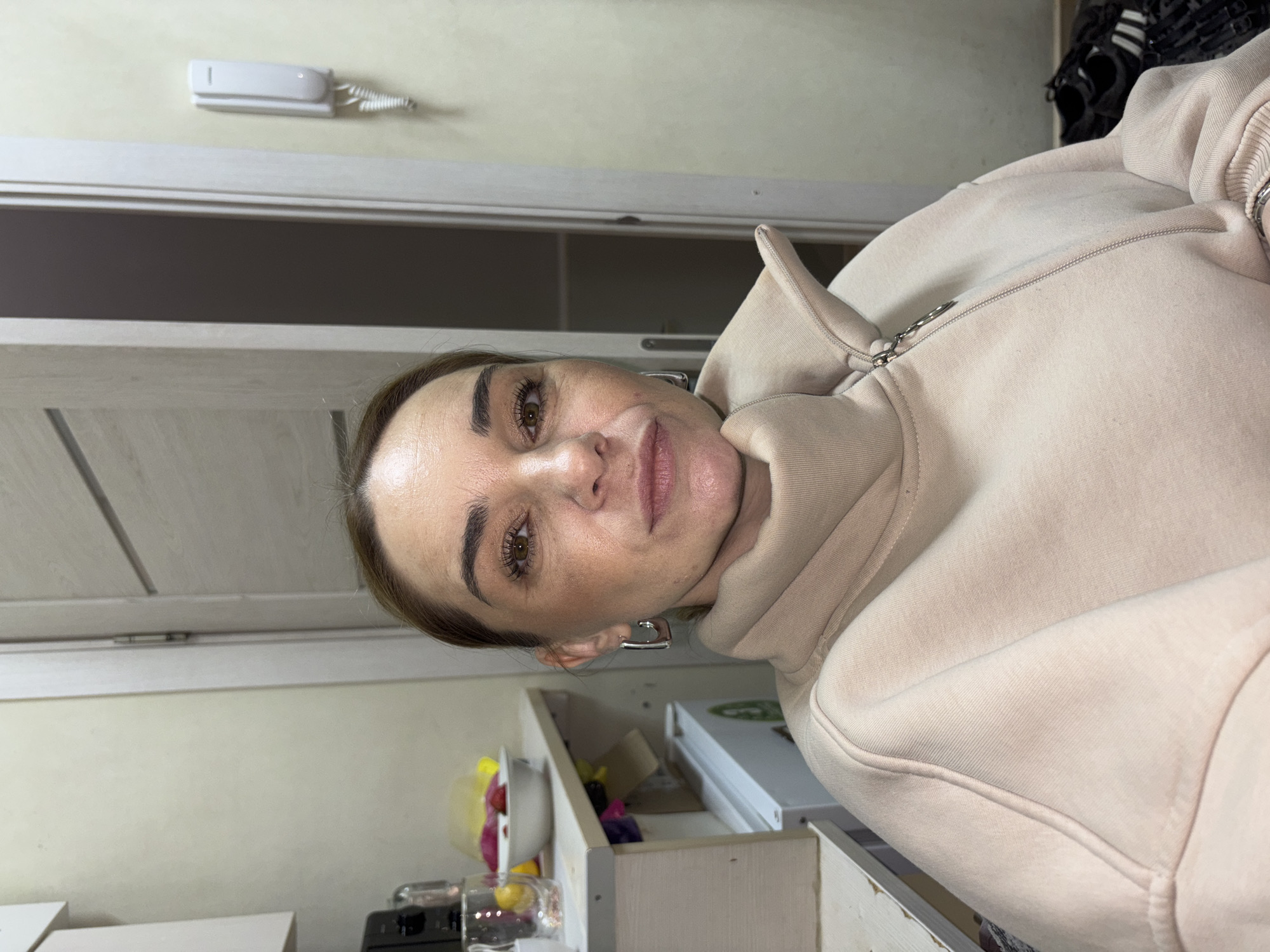 A woman is looking at the camera. She is wearing a tan hooded sweatshirt with a zipper. She has brown hair pulled back and brown eyes. She is wearing silver earrings. She appears to be inside a building, possibly a home, as there is a door in the background and a shelf with items on it. It is not possible to verify the location in Novoe Devyatkino, Russia from the image.