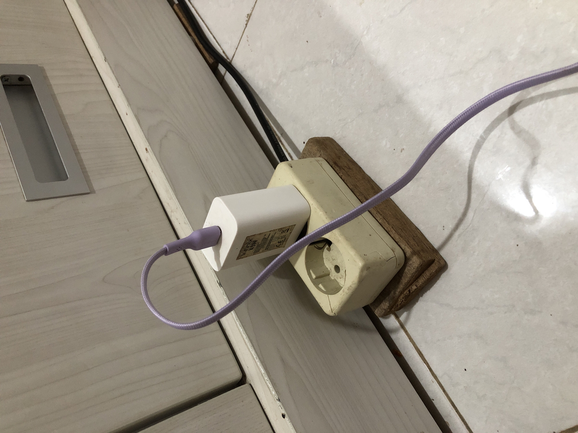 In a residential setting, possibly a bedroom or living area, in Pashiebeutong, Indonesia, a white Infinix 45W charger adapter is plugged into a beige, older model electrical socket. The socket itself is perched precariously on a small, rough wooden block. A thin, purple braided charging cable emerges from the adapter and snakes downwards, casting a shadow on the tiled floor. The background consists of a light-colored, textured wall, possibly part of a cabinet or furniture, with a silver drawer pull visible at the top. Black electrical cords are also visible behind the socket, suggesting other appliances or power sources. The overall impression is one of a functional, albeit somewhat makeshift, charging setup.