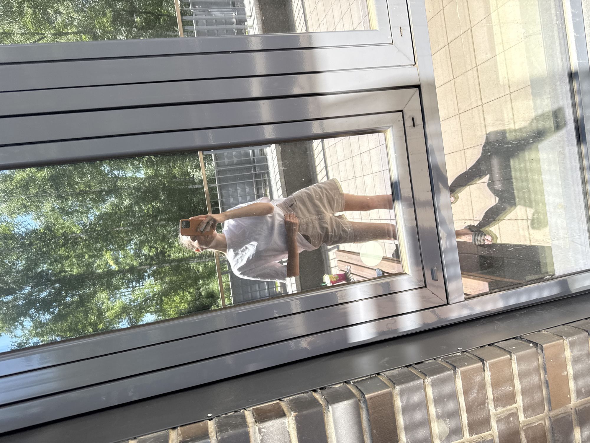 The image shows a reflection of a young woman in a window. She is standing on a balcony or patio, holding a smartphone up to take the photo. She is wearing a light-colored t-shirt and beige shorts. In the reflection, a table and a colorful drink are visible behind her. The background shows trees and a blue sky, suggesting an outdoor setting on a sunny day. The foreground shows a brick wall on the left and the window frame. The reflection also shows the lower half of her body, including her feet in sandals. The overall impression is a casual snapshot taken by the woman herself. Without further information or context, it is impossible to determine the specific location in Krasnoyarsk, Russia.
