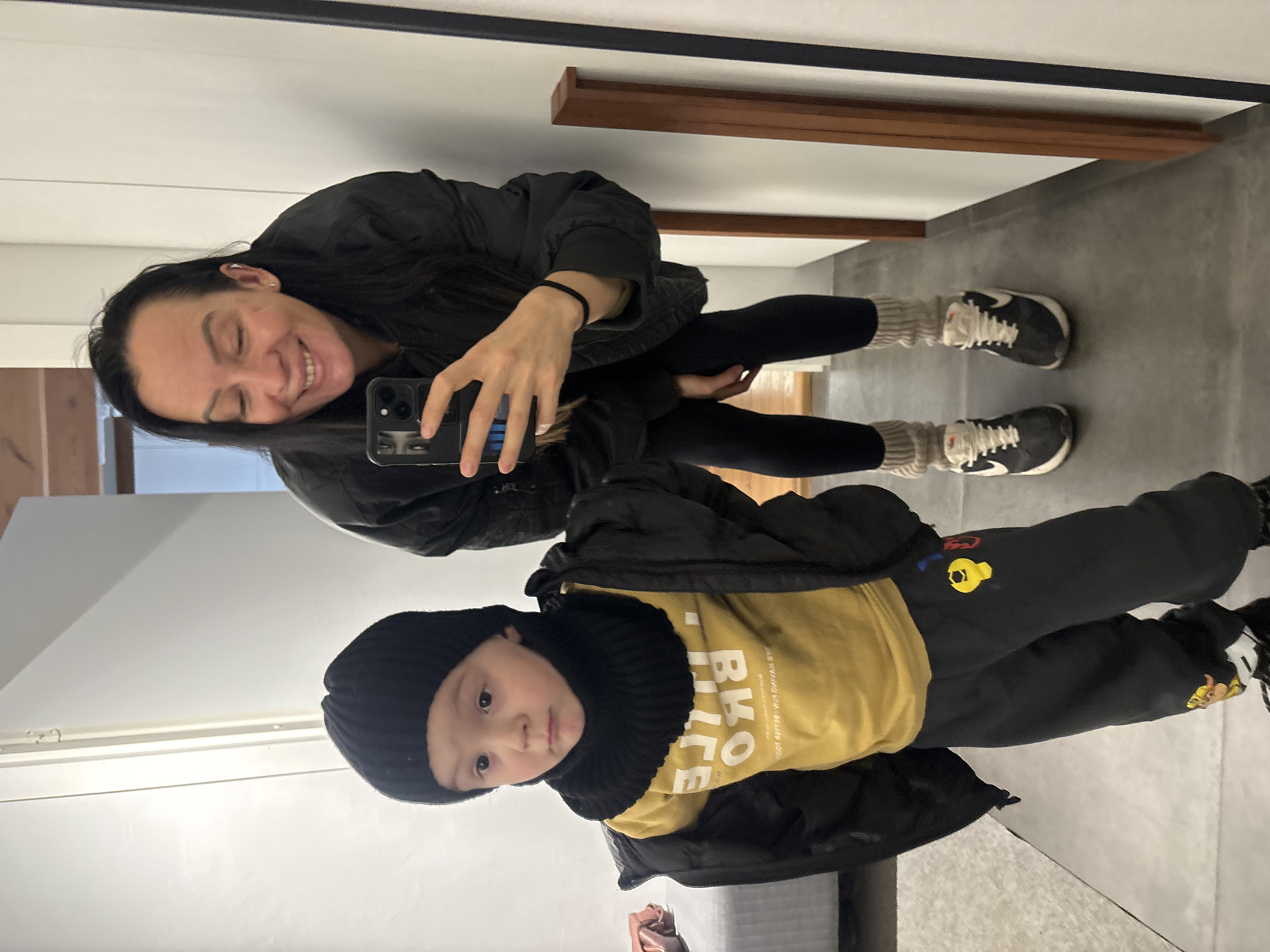 A woman and a child are posing in a mirror selfie indoors. The woman is wearing a black jacket, black leggings, and grey leg warmers with black and white sneakers. She is holding a phone in her right hand to take the picture and smiling. The child is wearing a black hat, a black scarf/neck warmer, a yellow t-shirt, and black pants with colorful cartoon-like patches, also wearing black and white sneakers. The floor appears to be gray tile or concrete. There's a brown wooden shelf or frame detail on the wall near the ceiling. The exact location in the city of Chaikovsky, Russia, cannot be determined from the image.