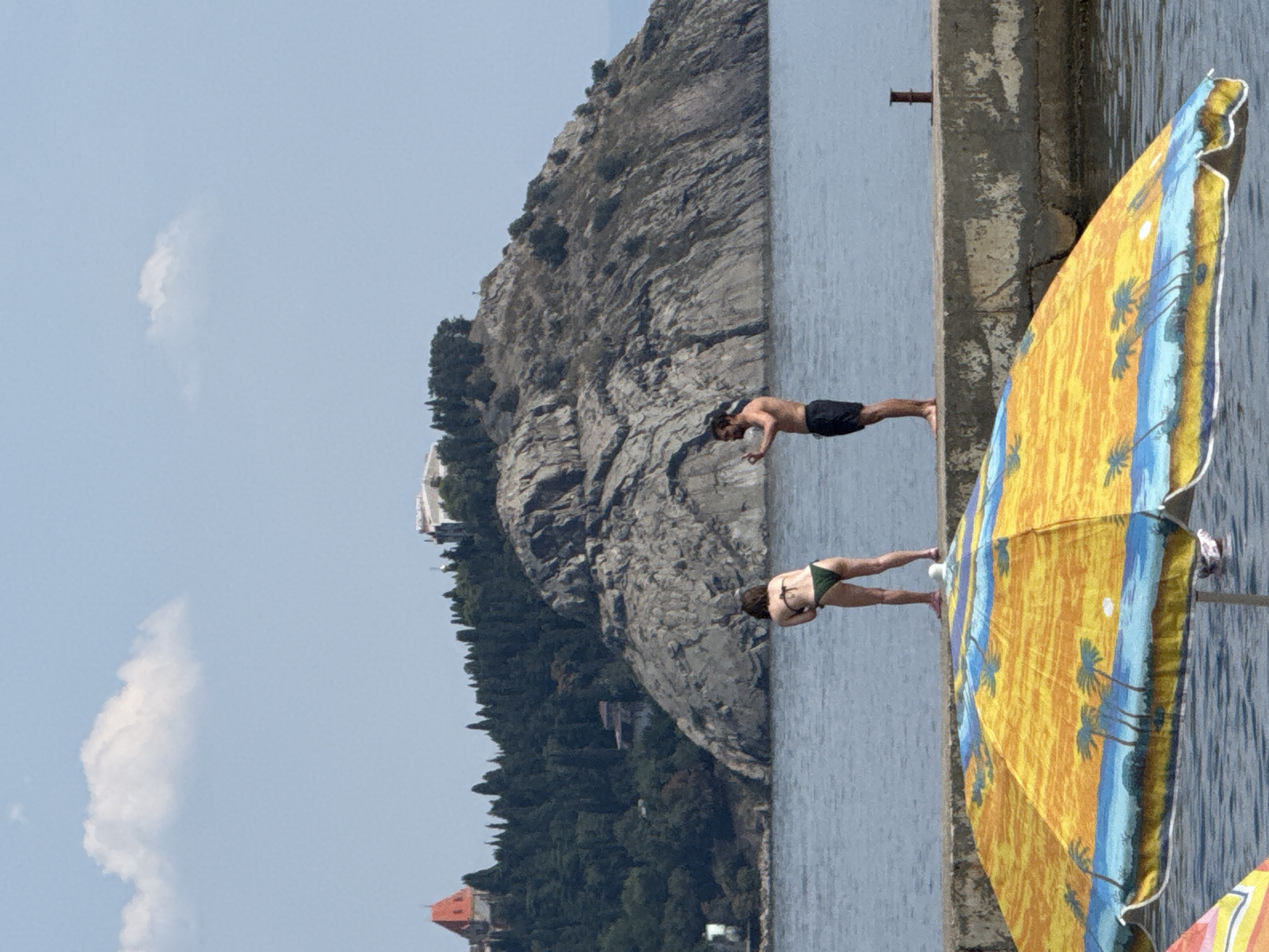 In the city of Partenit, Ukraine, a couple stands on a concrete pier overlooking a body of water. The woman, in a green bikini, is facing away from the camera, bending slightly. The man, wearing black swim trunks, is also facing away, holding a phone to take a photo. In the foreground, a colorful beach umbrella with a palm tree motif is unfurled, partially obscuring the water. Behind the couple, a rocky hill covered in dense green trees rises from the water. On top of the hill, a building with a red roof and other structures are visible. The sky above is a pale blue with a single white cloud. The overall scene depicts a sunny day at a coastal location.