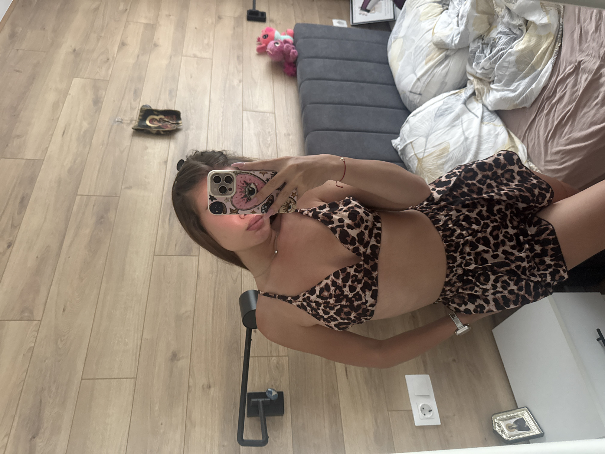 The image shows a woman taking a selfie indoors. She is wearing a leopard-print bra and shorts set. Her location appears to be a bedroom, evidenced by the bed with rumpled bedding in the background. Other elements visible include a gray padded headboard, wooden flooring, a small decorative item, and a power outlet. There is no information about the city of Unknown.