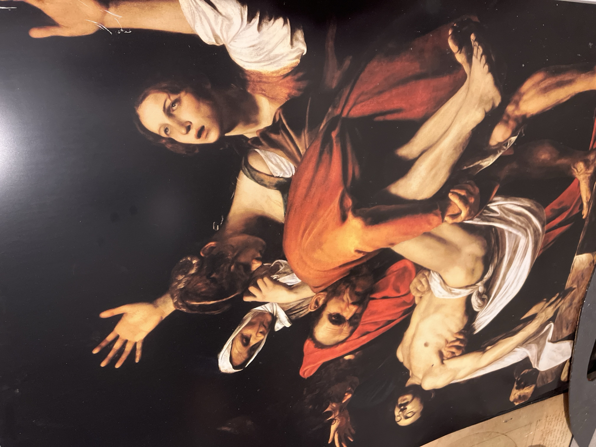 This image displays a photographic reproduction of Caravaggio's painting "The Entombment of Christ." The scene is composed of six figures arranged in a dramatic, triangular composition against a dark, unadorned background, rendered with strong chiaroscuro lighting that highlights the figures and their expressions.
At the focal point, the deceased, pale body of Jesus Christ lies horizontally, clad only in a white loincloth that wraps around his lower torso and legs. His head is tilted back, eyes closed, and his right arm hangs limply. He is being lowered onto a visible stone slab, presumably the entrance to a tomb, positioned at the bottom right.
Two male figures support Christ's body. On the right, a bearded, older man, wearing a vibrant orange-red tunic, stoops with a strained facial expression, holding Christ's legs and lower body. His bare, muscular right arm is prominent. To the left, a younger man with dark, flowing hair supports Christ's upper body and left arm, looking down with a somber gaze.
Behind these men, three female figures are depicted in various states of grief. An older woman with a white head covering stands to the left, looking downwards with a deeply sorrowful expression. Adjacent to her, a younger woman with dark hair bows her head, her hands clasped together near her chest, indicating intense sorrow. Further to the right, a third younger woman gazes upwards with an open mouth and wide eyes, her right arm dramatically extended towards the top right of the frame, conveying extreme anguish. She wears a white sleeve under an earth-toned garment.
The painting's environment is devoid of specific architectural or landscape details, intensifying the focus on the figures and their emotional interaction. The light source appears to be from the upper left, casting deep shadows and modeling the forms with stark clarity. This photographic reproduction is located in the city of Santeramo in Colle, Italy.