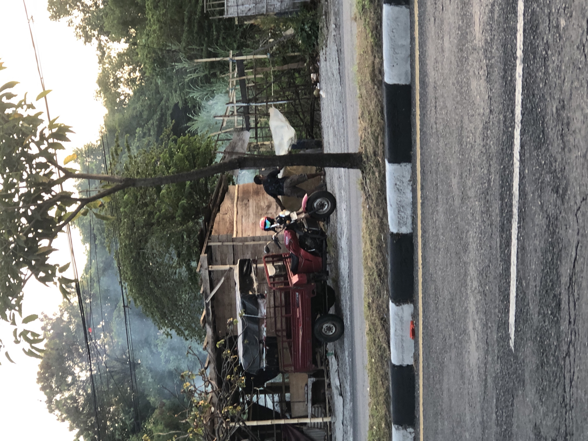 In Siring, Indonesia, a red three-wheeled motorcycle with a cargo bed is parked on a dusty, unpaved area beside a road. A person wearing a red helmet and a blue face shield is seated on the motorcycle, while another person in a dark shirt and khaki shorts is walking away from it, carrying a white sheet or tarp. Behind them are wooden structures, some appearing dilapidated and others constructed with bamboo, suggesting an informal settlement or construction site. In the background, trees and vegetation are abundant, with a hazy glow suggesting either smoke or the setting sun. The road in the foreground has a black and white painted curb and yellow and white lane markings.