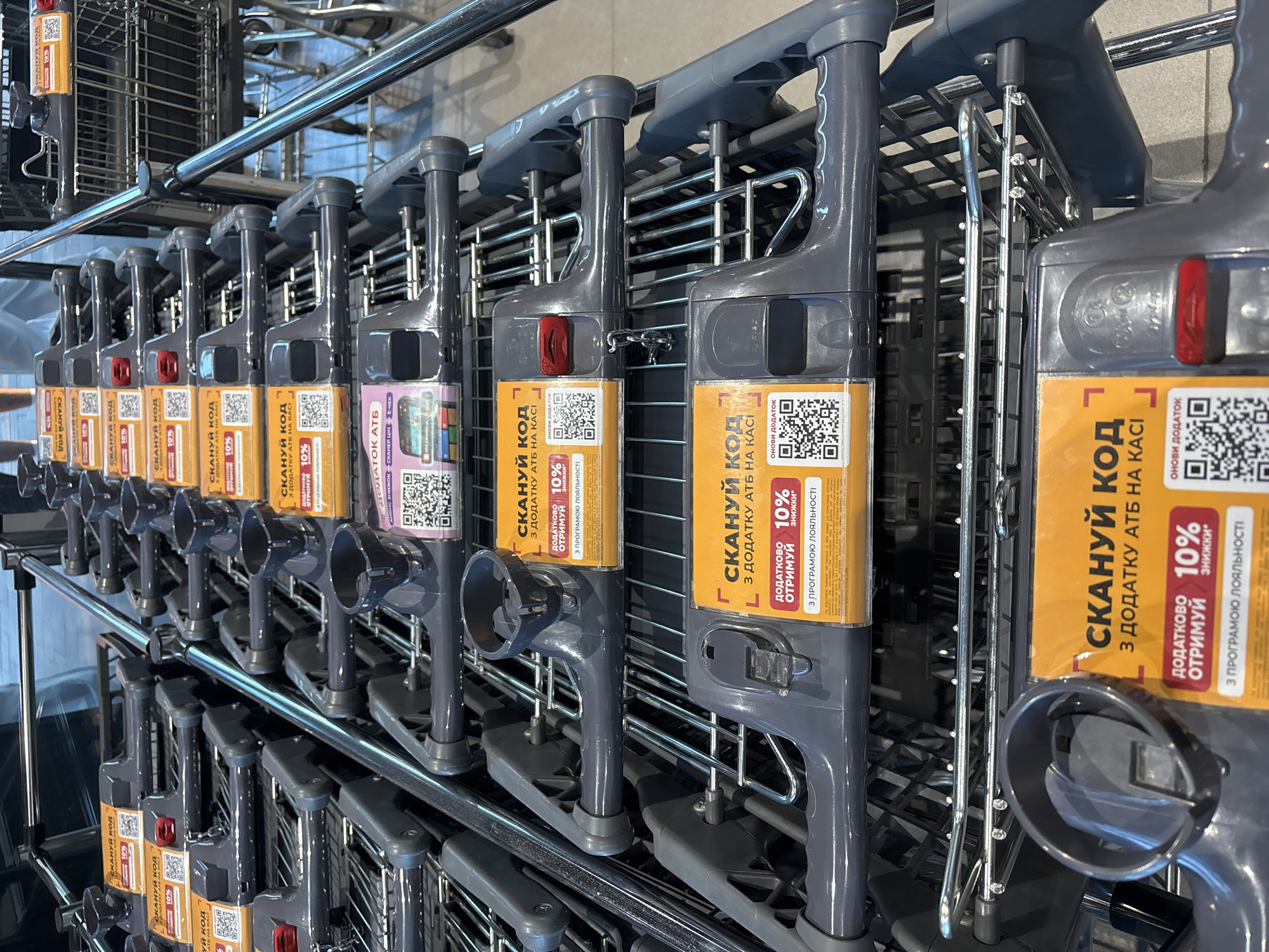 The image shows a row of shopping carts in what appears to be a supermarket. The carts are stacked in a line, with their handles facing forward. Some of the carts have yellow stickers with Ukrainian text and QR codes. The environment is indoors, with what looks like fluorescent lighting. The image is taken from a slightly elevated perspective, looking down at the shopping carts. The city of Bila Tserkva, Ukraine is mentioned, but there is no visual information in the image to confirm this location. There are no people visible in the image.