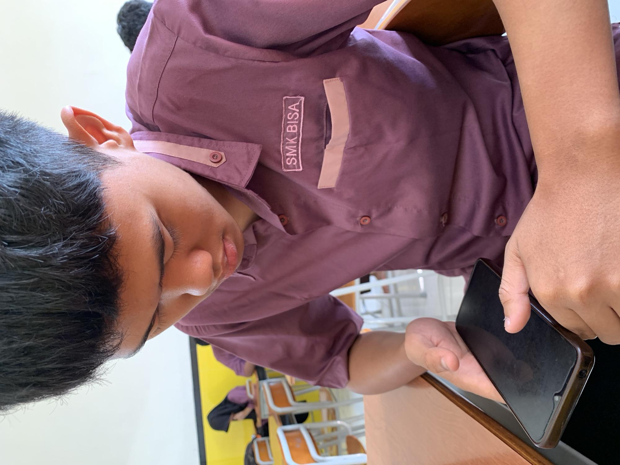 A young Indonesian male, wearing a maroon collared shirt with a patch that reads "SMK BISA" and a light pink pocket, is seated at a wooden desk in a classroom. He is looking down at a black smartphone in his hands. The background shows other students in maroon uniforms, some with headscarves, and desks with orange-trimmed chairs. A yellow stripe runs along the wall. The scene suggests a school setting in Rembun, Indonesia.