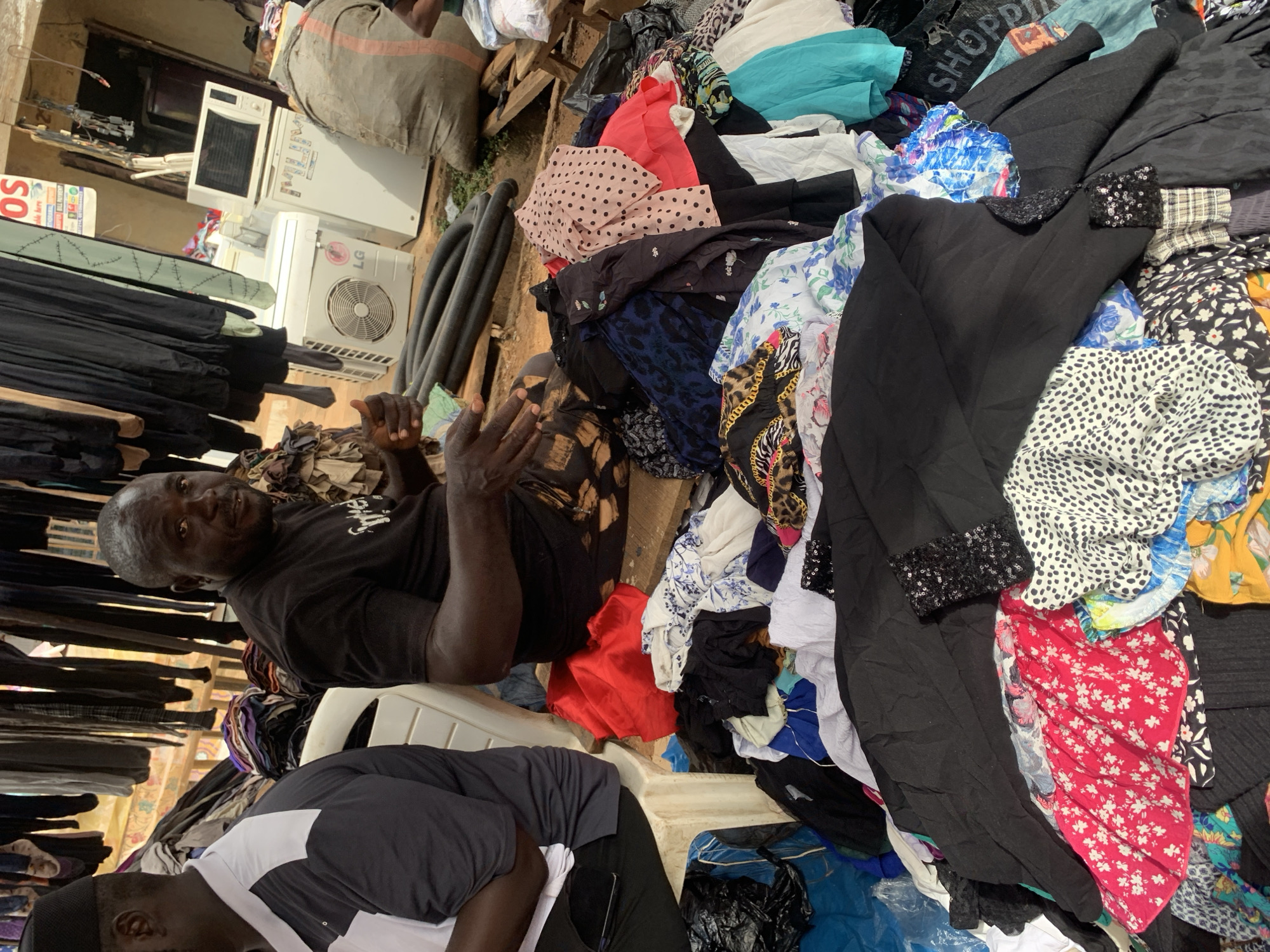 The image depicts an outdoor market scene in Nyanya, Nigeria, with two men present. In the foreground, a large pile of assorted clothing is displayed, featuring various patterns and colors including floral prints, polka dots, and solid black fabric. One man, wearing a black t-shirt and a patterned skirt, is seated on a wooden plank and gesturing with his hands. To his left, another man, wearing a black and white shirt and a black skullcap, is seated on a white plastic chair. Behind the clothing pile, several items of clothing hang on display racks. In the background, a white air conditioning unit, a refrigerator, and a microwave are visible, suggesting a mix of goods being sold. A large burlap sack is also present in the background, along with a sign that reads "OS." The ground appears to be dirt.