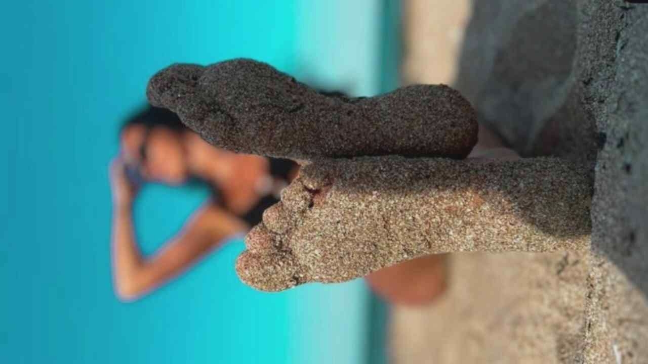 A low-angle, close-up shot focuses on two feet covered in sand, set against a blurred background of a person in a bikini on a beach. The feet are positioned in the foreground, with the toes of the left foot clearly visible and the sole of the right foot turned towards the viewer. The sand is granular and clings to the skin, creating a textured appearance. In the mid-ground, a person is visible from the shoulders up, their face obscured by sunglasses and their arm raised to shield their eyes from the sun. They are wearing a dark bikini. The background is dominated by a bright blue sky, with a hint of the ocean or a body of water at the horizon. The scene appears to be a beach setting, likely during daylight.