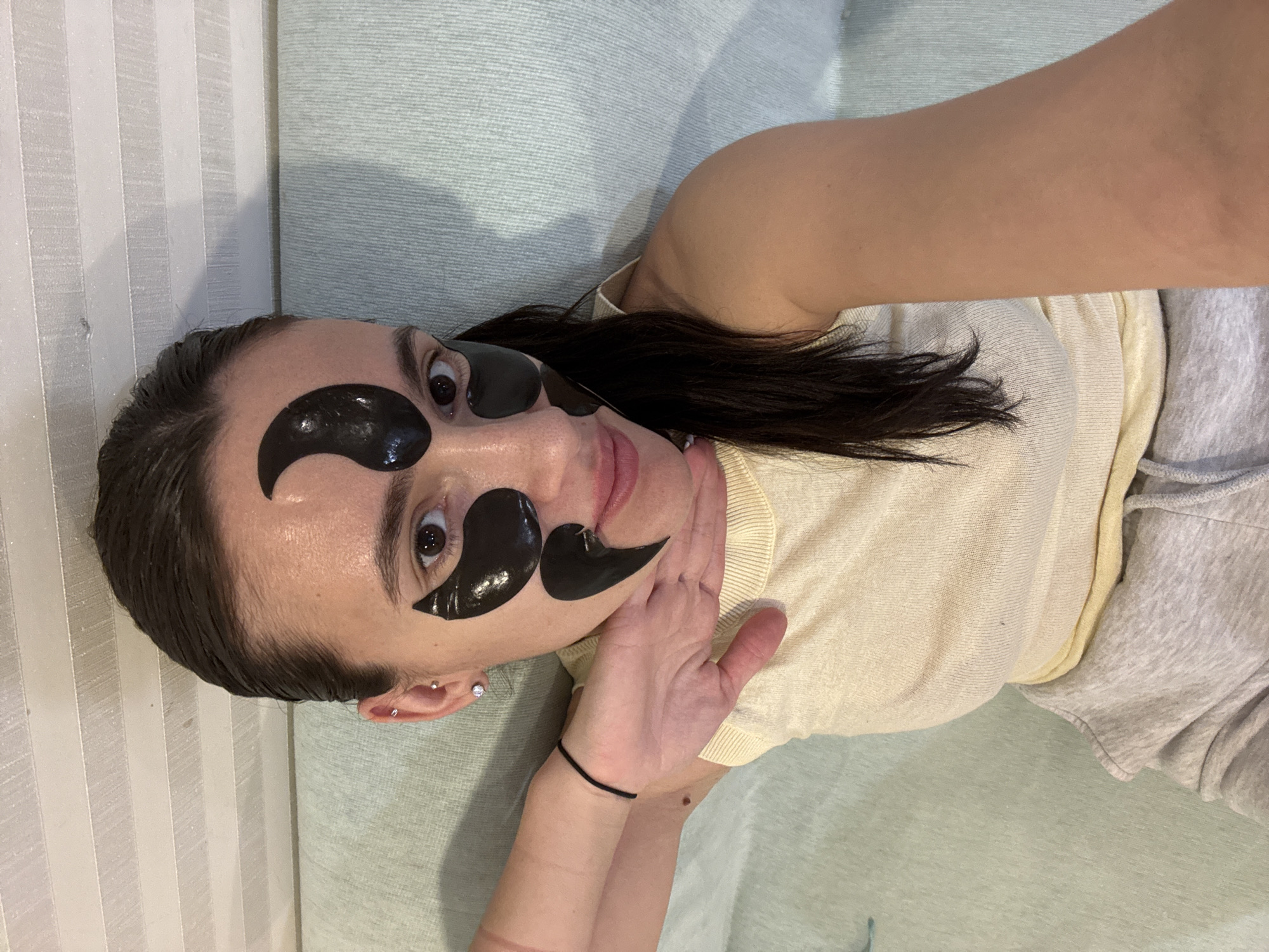 A woman is lying on a light blue textured surface, possibly a couch, wearing black under-eye patches and a light yellow top with grey sweatpants. Her head is tilted slightly to the left. She has dark hair, fair skin, and her eyes are open. She is holding her left hand near her throat. There is a white striped pattern visible in the top left corner. The location is not discernible from the image.