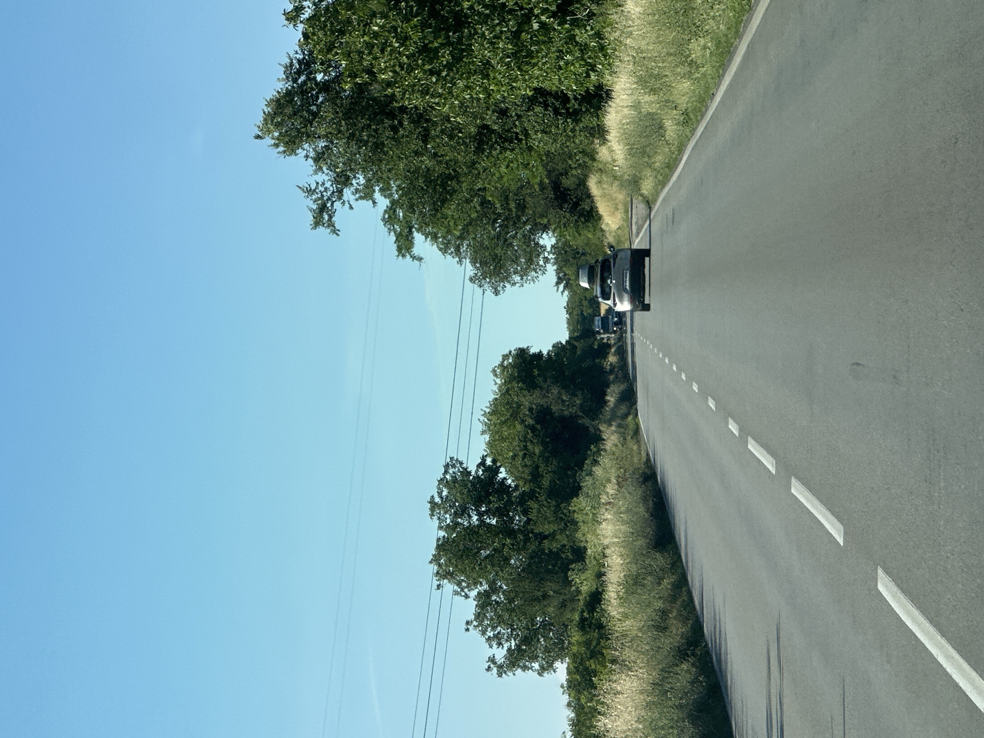 The image depicts a two-lane asphalt road in a rural area, likely Samovodene, Bulgaria, under a clear blue sky. The road has dashed white lines marking the lanes. On the right side of the road, a dense line of green trees borders the asphalt, with tall, dry grass in the foreground. On the left side, a similar arrangement of tall, dry grass is followed by more trees and vegetation, with power lines visible above them. Two cars are driving away from the viewer down the road. The car closest to the viewer is a silver SUV with a cargo carrier on its roof. Further down the road, another vehicle, possibly a truck, is visible. The overall scene suggests a sunny day in a scenic, natural environment.