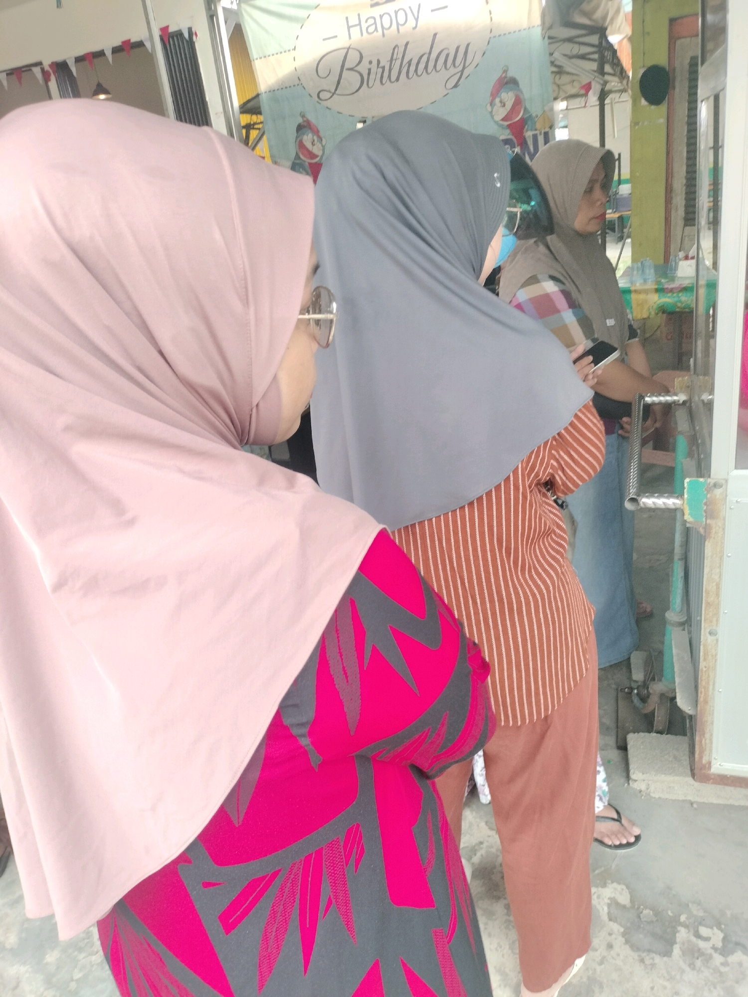 The image depicts three individuals in Pembataan, Indonesia, standing near what appears to be a food stall or shop entrance. The foreground is dominated by the back of a person wearing a light pink hijab and a dress with a striking magenta and dark gray floral pattern. To their right, a second person, partially visible, wears a gray hijab and an orange and white striped shirt, holding a mobile phone. The third person, further to the right and in the background, is a woman wearing a beige hijab and a patterned top, looking towards the right. A banner with "Happy Birthday" written in cursive and an illustration of what looks like Doraemon is visible in the background. The environment suggests an outdoor setting with a concrete floor and what looks like metal grating or a fence to the right.