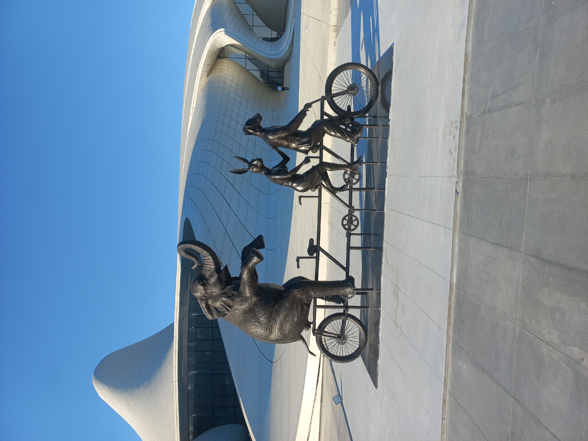 This image captures a bronze sculpture of three anthropomorphic animals on a tandem bicycle in front of the Heydar Aliyev Center in Baku, Azerbaijan. The sculpture depicts an elephant in the front, appearing to be pedaling, followed by a rabbit, and then a dog in a suit and tie. The background features the distinctive, wave-like architecture of the Heydar Aliyev Center, characterized by its white, flowing curves and large glass windows. The sky above is a clear, bright blue, indicating a sunny day. The ground in front of the sculpture is paved with light gray tiles, and a darker tiled area forms a base for the sculpture. The overall scene is modern and artistic, highlighting the unique architectural landmark of Baku.