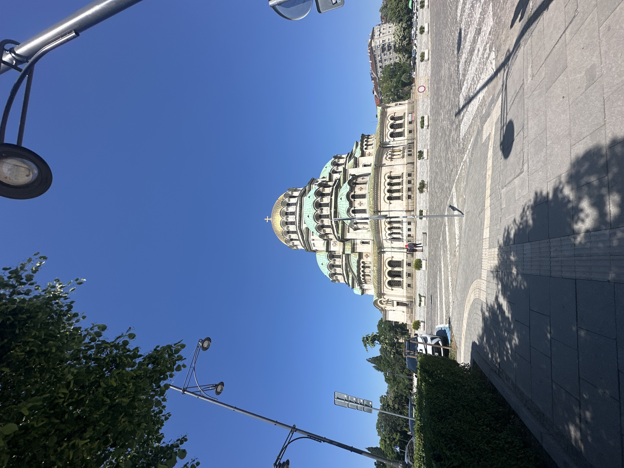 The image depicts the Alexander Nevsky Cathedral in Sofia, Bulgaria, under a clear blue sky. The cathedral's golden dome and intricate facade are prominently displayed. To the left, streetlights and foliage frame the view, while on the right, a wide pedestrian area with cobblestone patterns and shadows is visible. A few people can be seen in the background, along with parked cars and greenery. The composition emphasizes the cathedral's architectural grandeur and its urban setting.