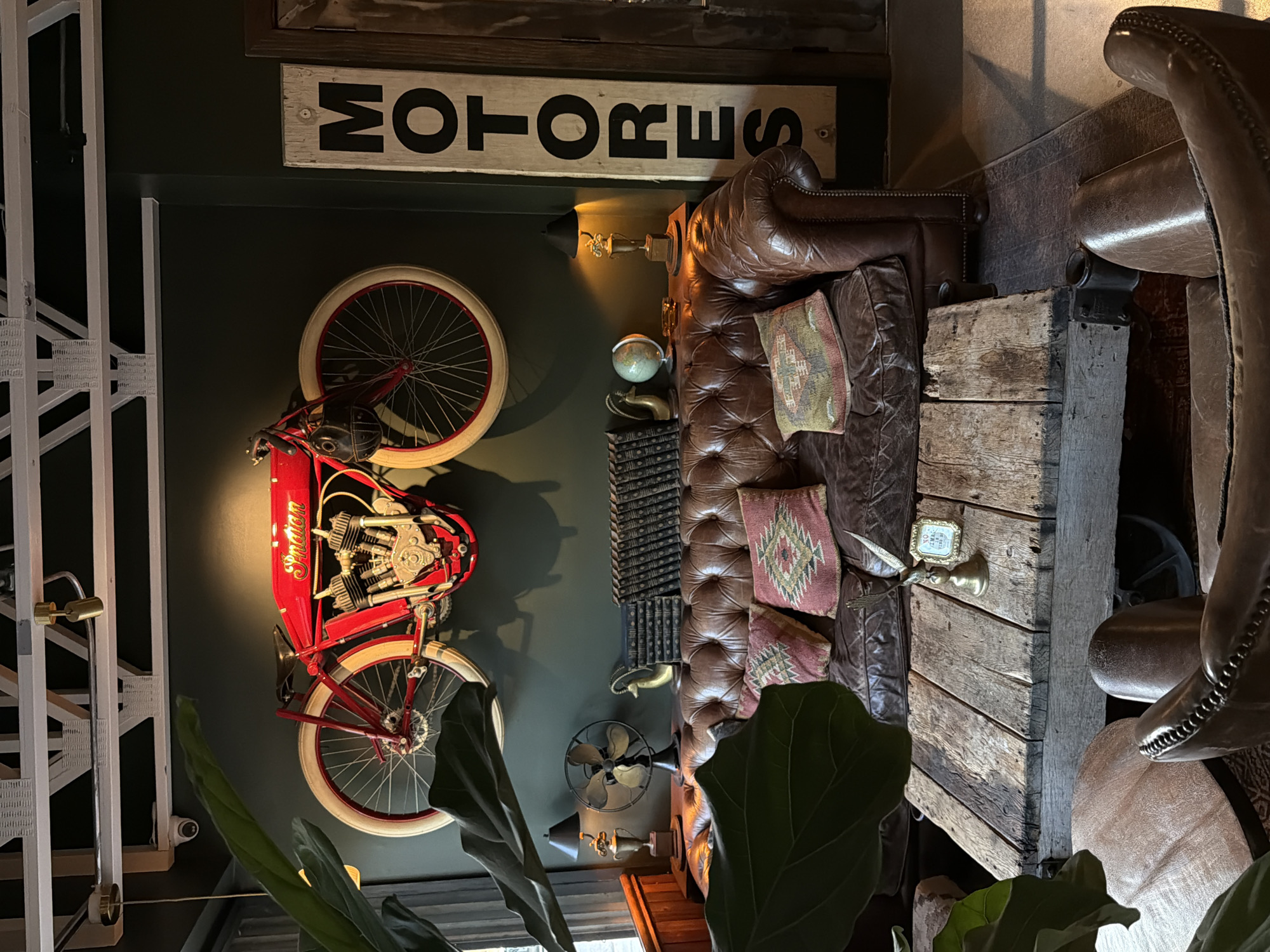 The image presents an interior design scheme with a vintage or industrial aesthetic, photographed at an angle.
The main elements include:
- A vintage-style motorcycle mounted on a dark-colored wall. The motorcycle is predominantly red with white rims and features the word "Indian" in gold lettering.
- Above the motorcycle, a sign with the word "MOTORES" in bold black letters.
- A leather couch with tufted cushions, adorned with decorative pillows.
- A rustic, wooden structure adjacent to the couch, which seems to serve as a side table or decorative element.
- Additional decorative items such as a globe, a small fan, books, and wall lamps are also present.
- There are indications of indoor plants in the foreground, obscuring part of the view.
The image suggests a bar, a club, or a restaurant in the city of West Hollywood, United States of America.