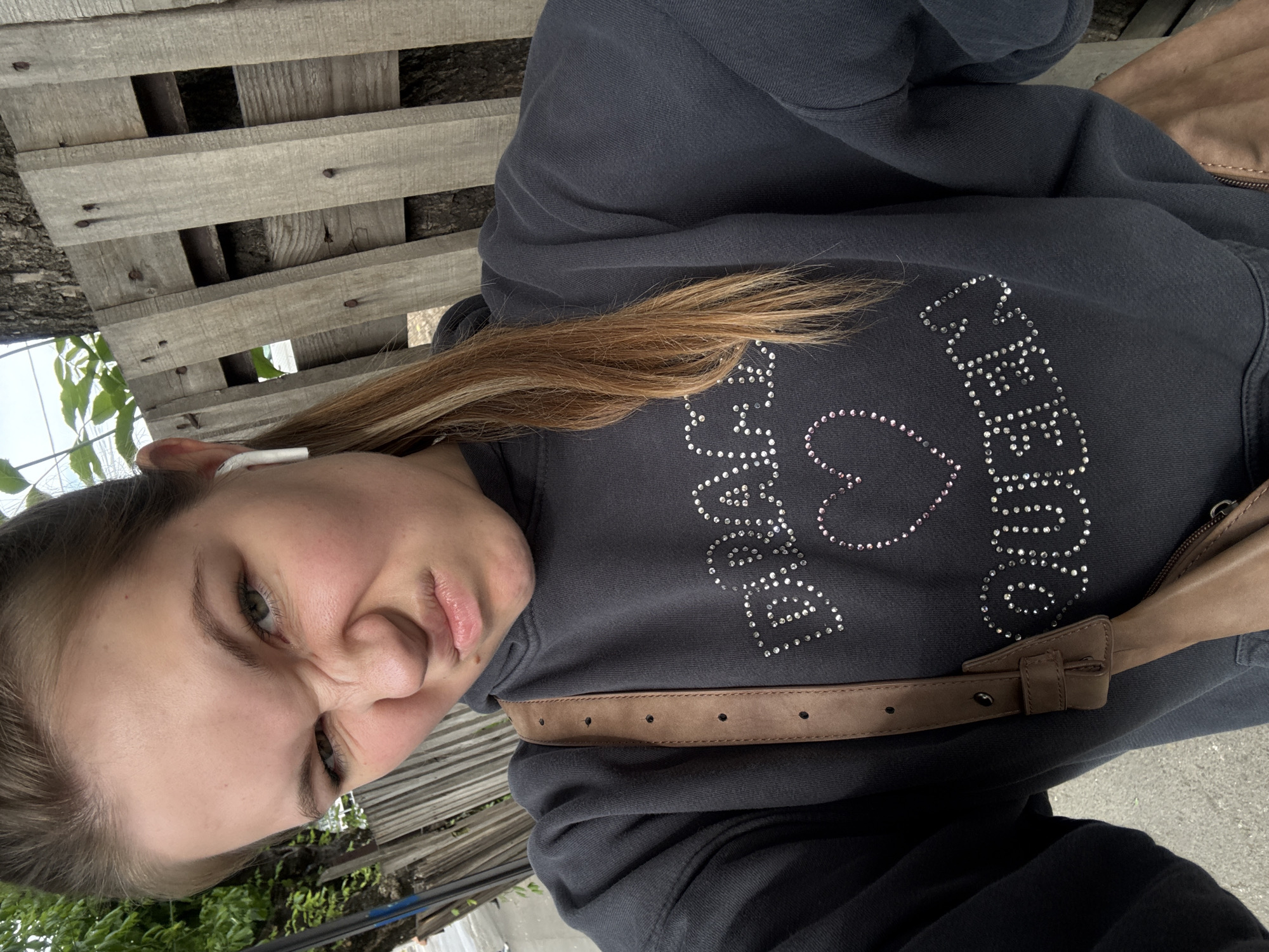 A young woman is sitting outside. She is wearing a dark-colored hoodie with rhinestone decorations spelling out "Peace Love Queen." A brown leather strap or belt is slung across her body. She has light skin, light brown hair with blonde highlights, and an earbud in her ear. She is making a scrunched-up face at the camera. She is sitting on a wooden pallet. Green foliage is visible in the background. Concrete ground can also be seen. The location is likely Vozdovac Urban Municipality, Serbia, but the image does not offer any definitive evidence to confirm.