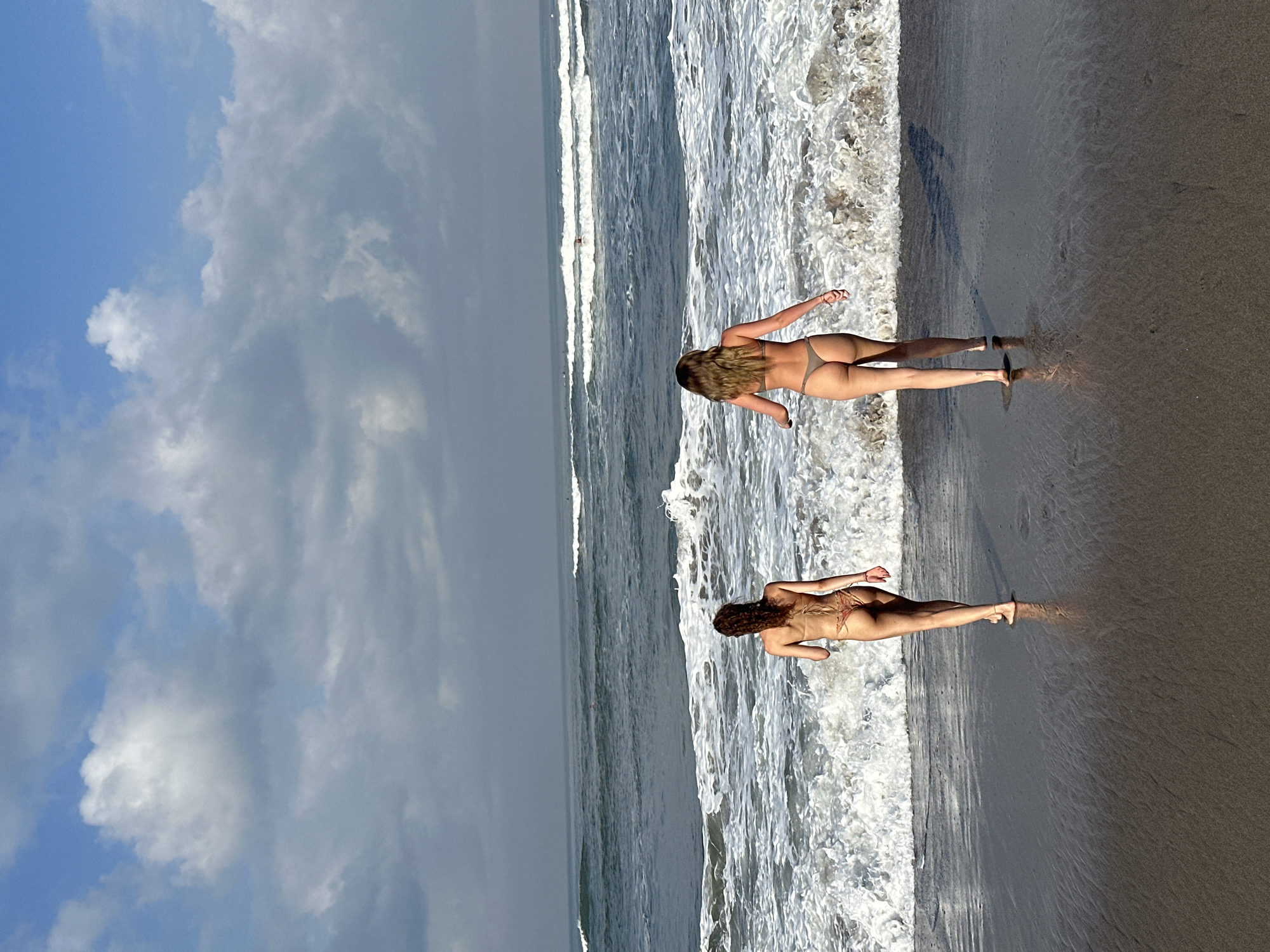 In North Kuta, Indonesia, two women in bikinis walk towards the ocean on a beach. The woman on the left has curly brown hair and is wearing a tan bikini. The woman on the right has blonde hair and is wearing a grey bikini. They are walking on wet sand with waves crashing in front of them. The ocean stretches out to the horizon under a cloudy blue sky.