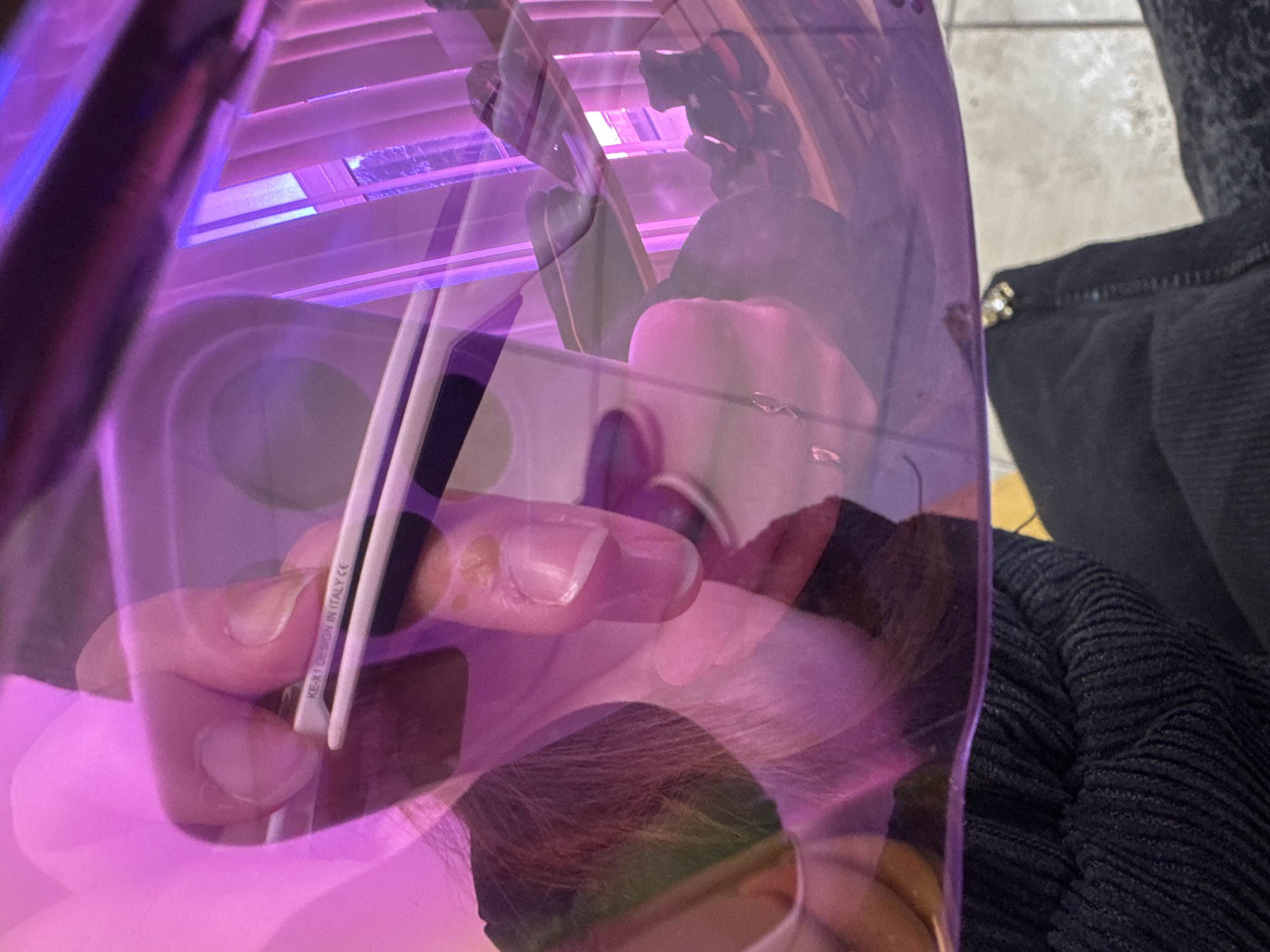 The image captures an indoor scene observed through a translucent, curved, purple-pink material, likely a visor or face shield, heavily tinting the visual information. In the mid-ground, a human hand is prominently featured, holding a pair of white-framed glasses. Inscriptions "KE-X1 DESIGN IN ITALY CE" are visible on the temple of the glasses. Behind and partially obscured by the hand, the blurred outline of a person's face, including hair and an ear, is visible, suggesting the perspective of the individual looking through or wearing the transparent object. The lower portion of the image shows dark, ribbed clothing on the left and dark trousers with a metallic zipper and embellishment on the right, resting above a light-colored tiled floor. In the background, visible through the filter, is a window equipped with vertical blinds, revealing a glimpse of outdoor foliage. Below the window, a shelf or rack holds several pairs of shoes, primarily dark with occasional red accents. The exact location within Прикамский, Russia, is not discernible from the provided visual data.