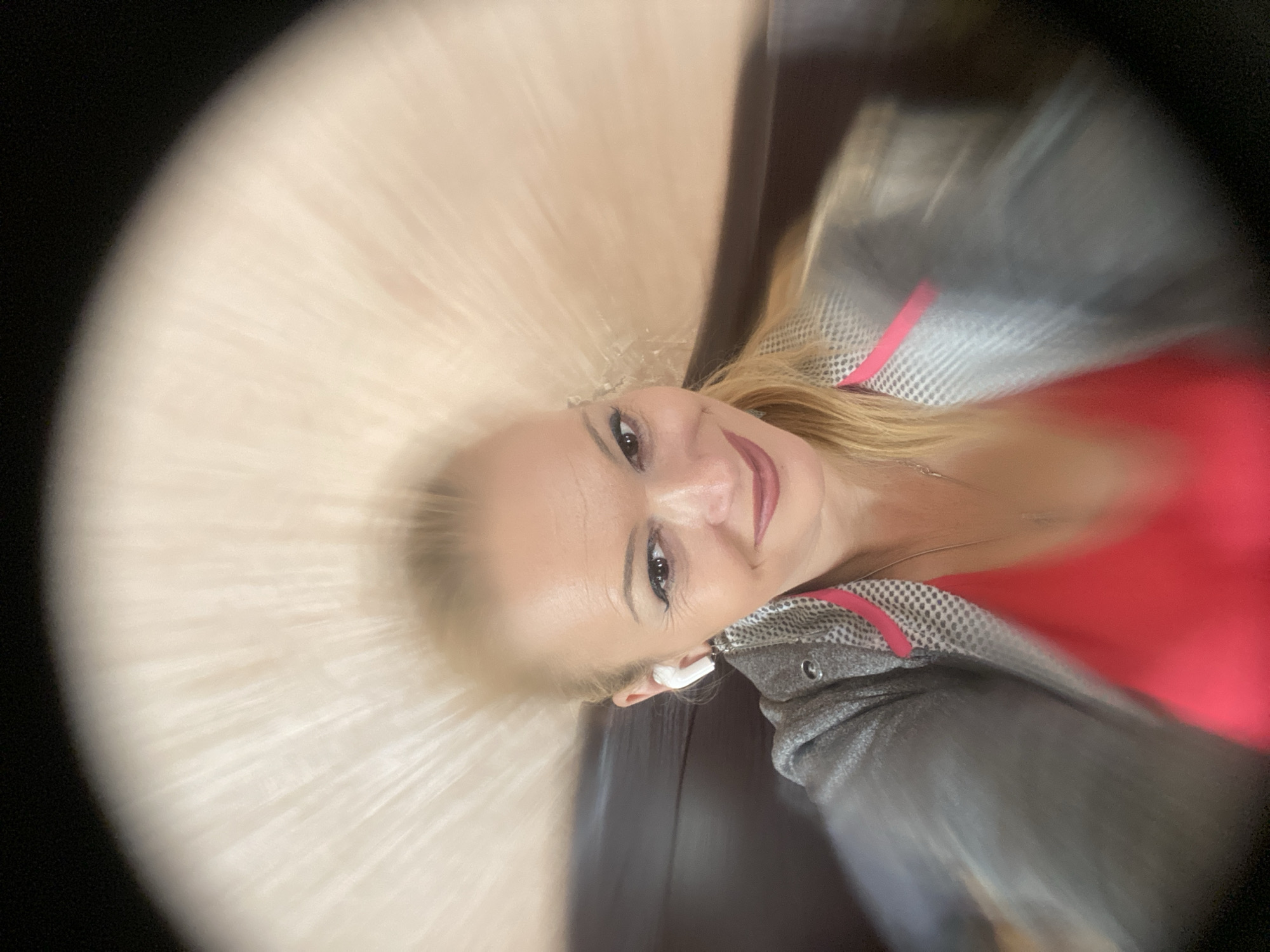 A woman is captured in a selfie, looking into the camera with a slight smile. She has blonde hair pulled back, styled away from her face. She is wearing white earbuds in her ears, a grey jacket with pink trim over a bright pink top, and a delicate silver necklace. The background is blurred, showing streaks of light radiating from behind her head, suggesting a bright, possibly artificial light source. The composition is a close-up shot, focusing on her face and upper body, with the edges of the frame darkening into a vignette. The image was taken indoors, possibly in a booth or seated area, indicated by the dark wooden surface visible to her left. The specific location in the city of Chernihiv, Ukraine, cannot be determined from the visual information present in the image.