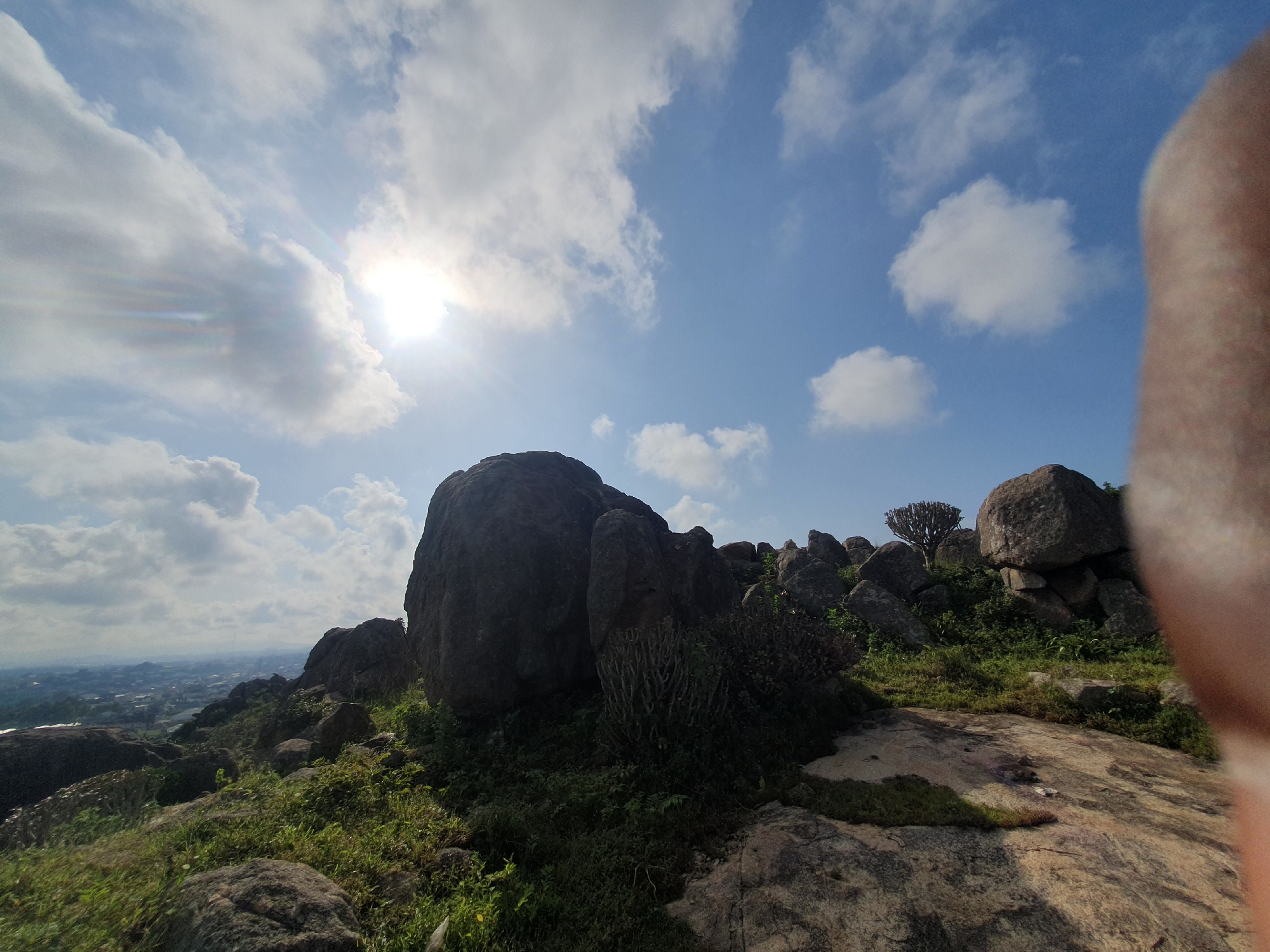 The image captures a panoramic view of Gura Top, Nigeria, characterized by a rocky, mountainous terrain under a bright, partly cloudy sky. The sun is visible, casting a bright glow through a break in the clouds on the left side of the frame, with some lens flare present. The foreground is dominated by large, dark, rugged boulders scattered across a grassy, uneven surface. Vegetation, including green shrubs and grass, grows between the rocks and on the slopes. In the middle ground, more rocky outcrops and scrubby vegetation are visible, leading towards a distant view of a city or town, with buildings and structures dotting the landscape. A single, small, gnarled tree stands on a rocky peak in the middle distance. A blurry, fleshy-toned object, likely a thumb or finger, is prominently in the right foreground, partially obscuring the view of the landscape. The overall impression is one of a natural, elevated landscape overlooking a populated area.