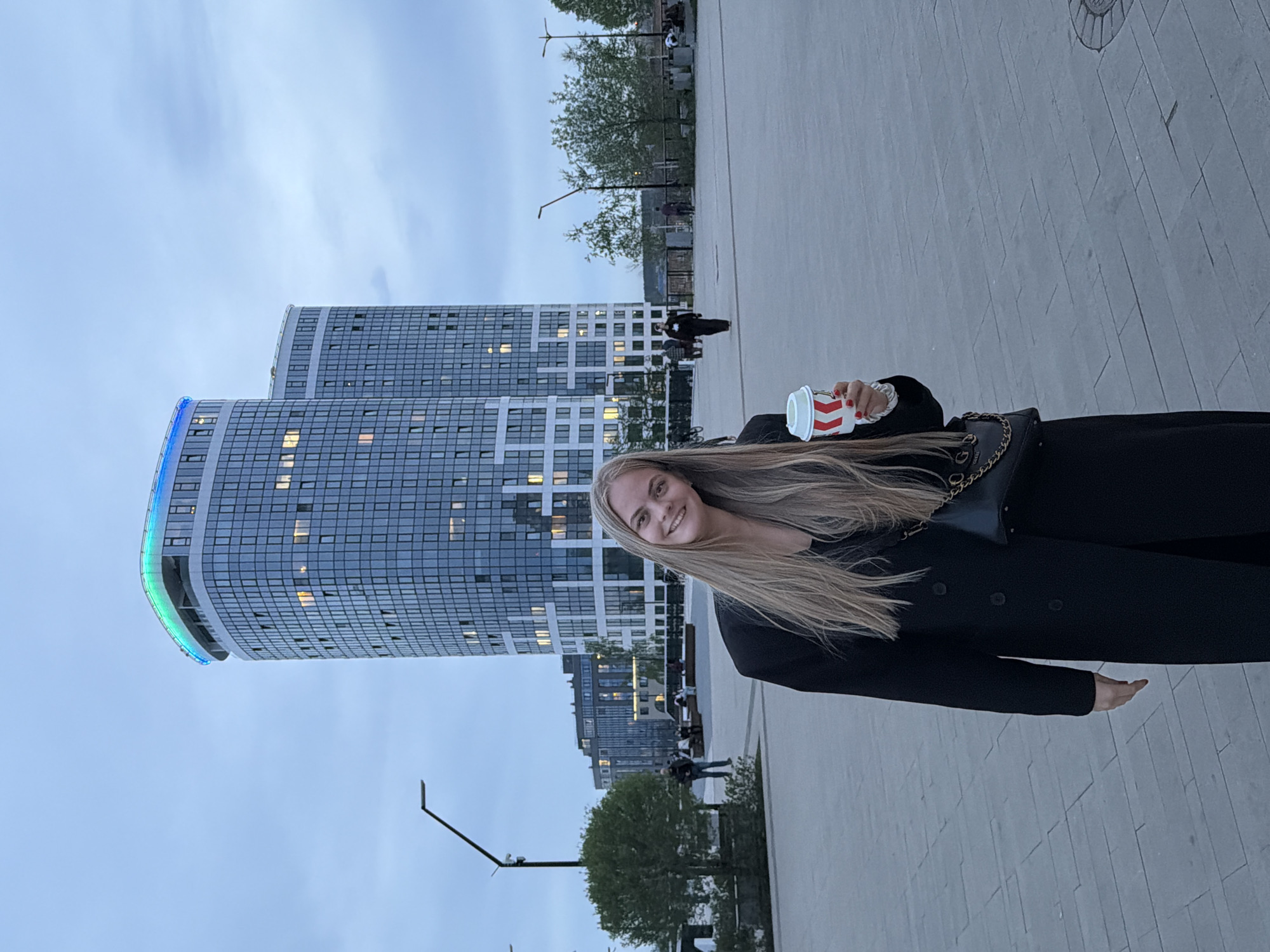 A woman is holding a drink while posing outside. The area appears to be a plaza. Behind her, there are modern buildings with many windows. Trees and street lamps are also visible in the background. The location is Izhevsk, Russia.