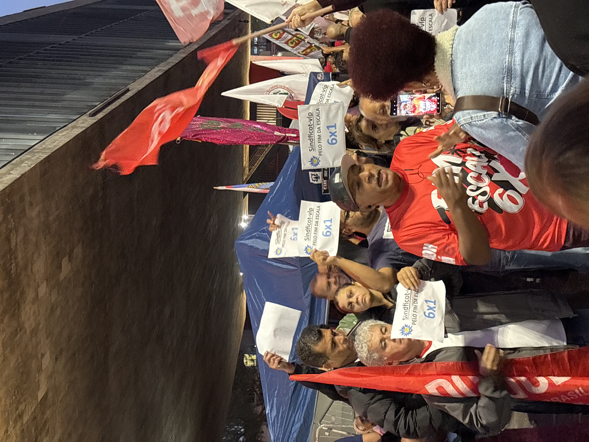 The image shows a group of people gathered in what appears to be a protest or demonstration. Many are holding signs with the text "Sindficot-vlp PELO FIM DA ESCALA 6x1". Some people are holding red flags. One person is taking a picture of a man in a red t-shirt with a cell phone. The crowd appears to be under a bridge or large structure. The banners and signs suggest the event is likely related to labor or workers' rights, specifically calling for the end of the "6x1" scale, possibly referring to a work schedule or condition. The location of São Paulo, Brazil is written on the flag.