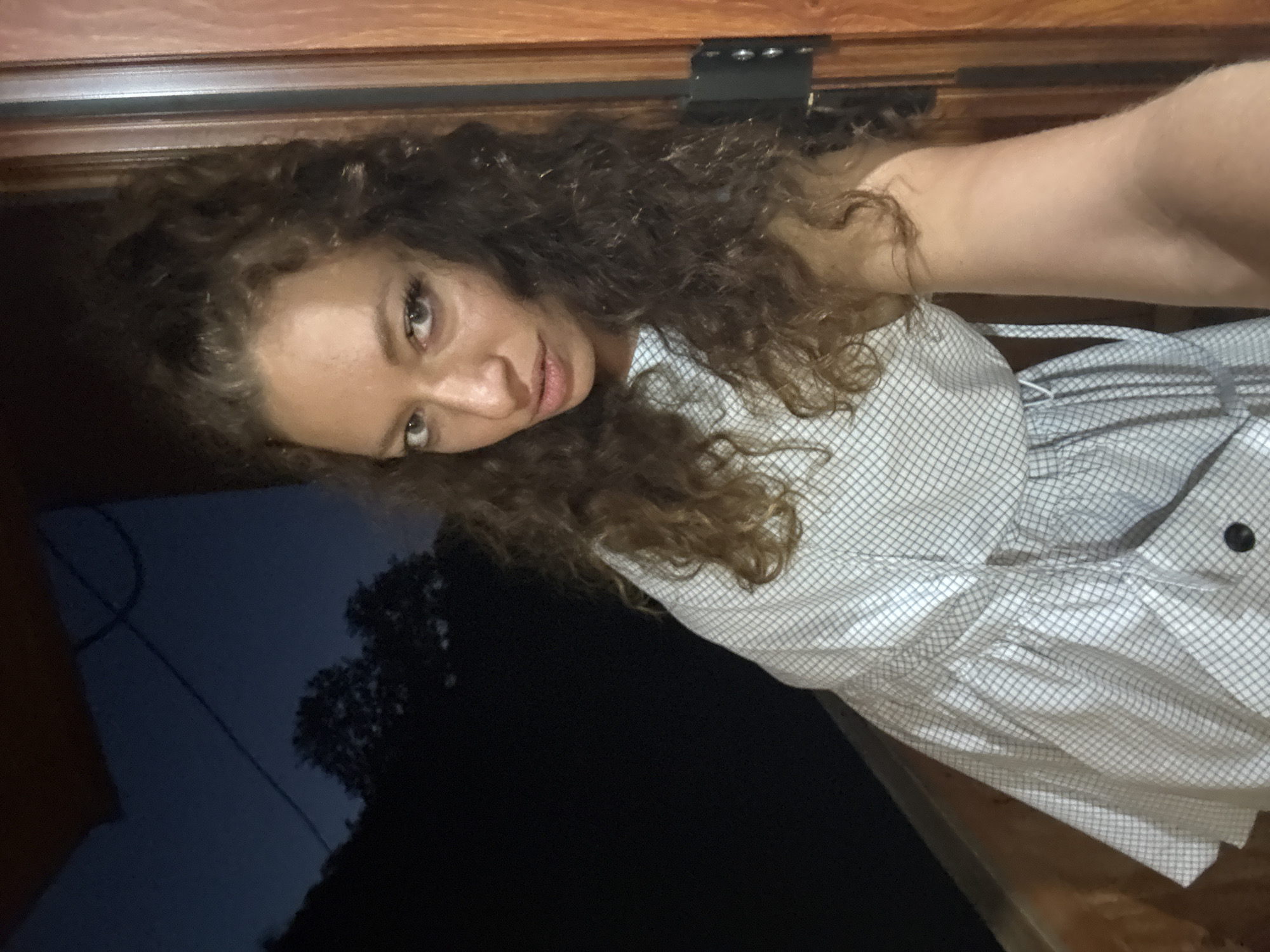 A woman with curly brown hair is facing the camera. She is wearing a white and gray patterned dress with a ruffled skirt. She is indoors, standing near a dark window with a glimpse of trees and sky visible outside. The background is dimly lit. The location appears to be Bắc Hà, Vietnam.