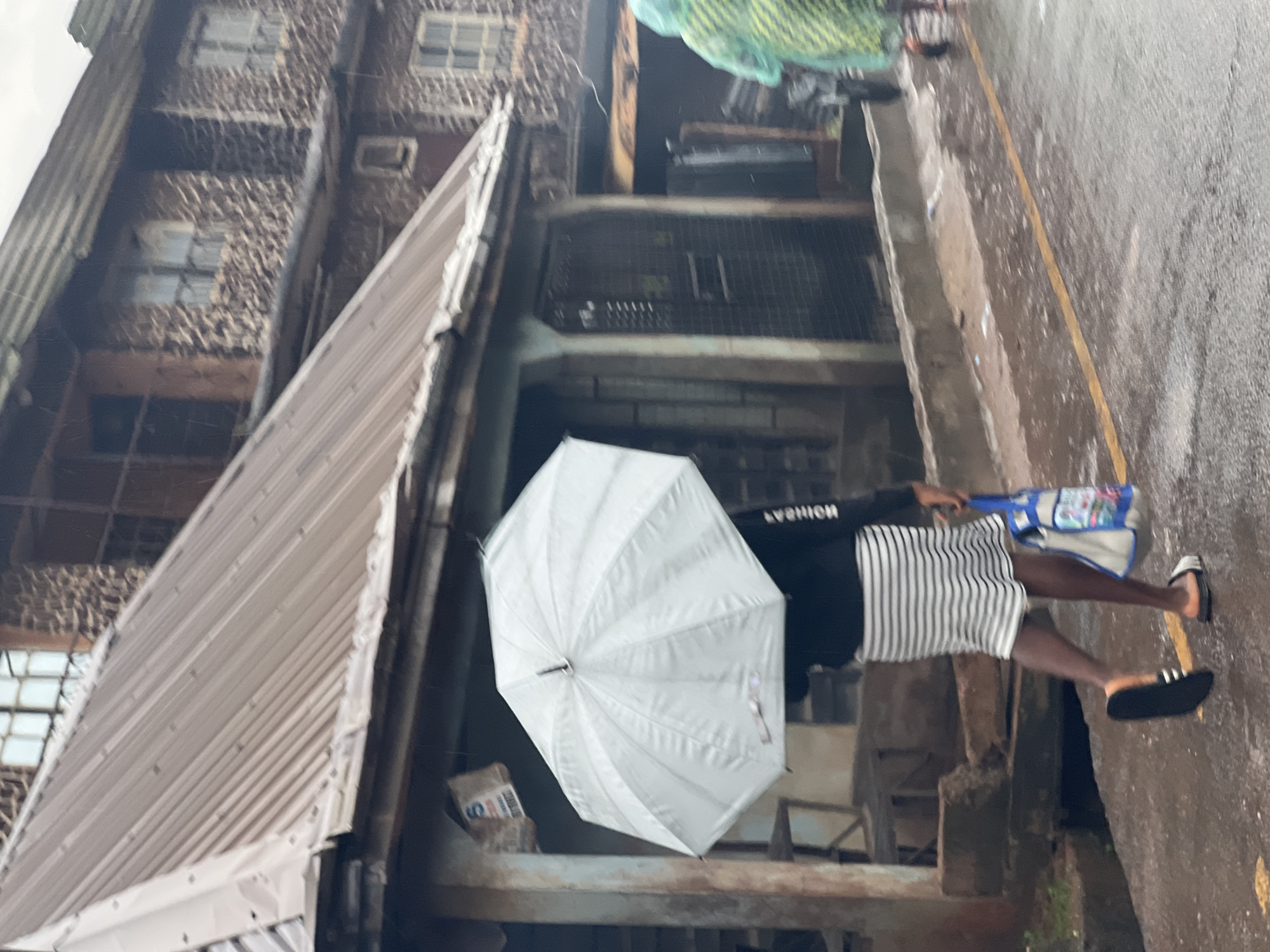 The image shows a person holding a white umbrella in what appears to be a rainy day in Nkpor, Nigeria. The person is wearing a black long-sleeved shirt and a black and white striped skirt. They are also carrying a blue and white bag. The background shows a weathered building with stone walls and corrugated metal roofing. The ground is wet, indicating recent rain. There is a yellow line painted on the ground, suggesting a roadside or pathway. The lighting is diffused, consistent with overcast or rainy weather. The overall impression is one of a mundane moment captured during inclement weather. No visible text is discernible in the image.