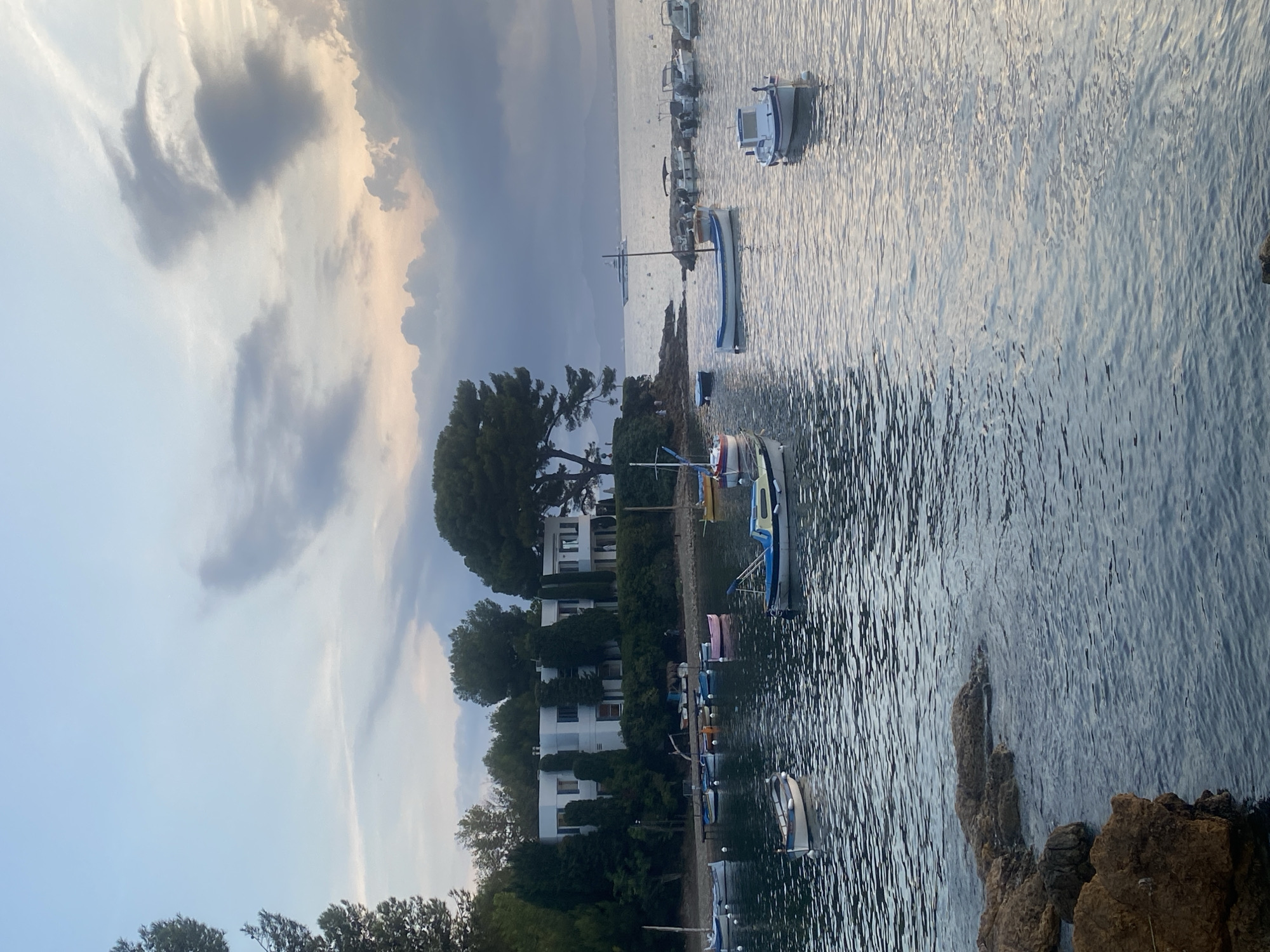 The image depicts a serene waterfront scene in Antibes, France, likely during the late afternoon or early evening, given the soft, diffused light and the dusky sky. In the foreground, weathered, reddish-brown rocks are partially submerged in calm, rippling water. Beyond the rocks, numerous small boats are anchored in a bay. These boats vary in size and design, with some being traditional wooden vessels and others more modern recreational craft. Several are painted white with blue accents, while at least one is a striking pink.
Across the water, a modern, multi-story white building with clean lines and numerous windows is nestled amongst lush green trees. The architecture suggests a private residence or a small hotel. The building's facade is partially obscured by the dense foliage, with tall, slender trees and broader, rounded evergreens.
In the middle ground, a larger, more distant vessel, possibly a yacht, is visible on the horizon, adding a touch of scale and luxury to the seascape. The sky above is a canvas of soft blues and grays, with dramatic, wispy clouds tinged with pastel shades of pink and orange from the setting sun. The overall impression is one of tranquility and Mediterranean charm. There are no discernible people in the immediate foreground or engaged in any specific actions within the frame. The scene is static and peaceful.
