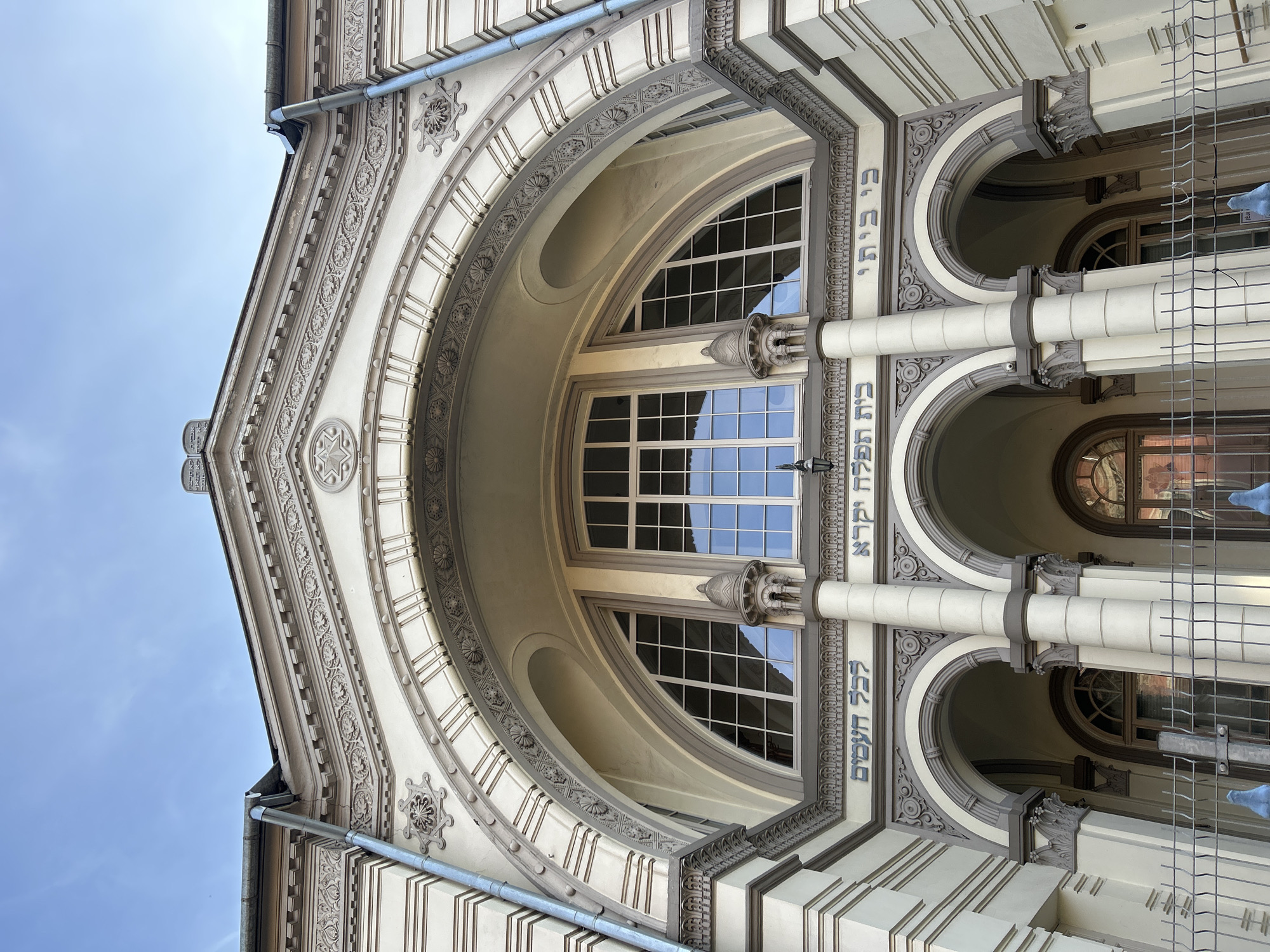 The image shows the exterior of the Choral Synagogue of Vilnius, Lithuania, a historical building with a light beige facade and ornate architectural details. The upper portion of the building features a large arched window with multiple panes, set within a decorative archway. Above the archway, Hebrew inscriptions are visible, along with decorative moldings and a Star of David emblem. The lower part of the facade displays a series of smaller arched entrances, supported by columns. A metal fence is present in the foreground, obscuring a clear view of the ground level. The sky is clear and blue.