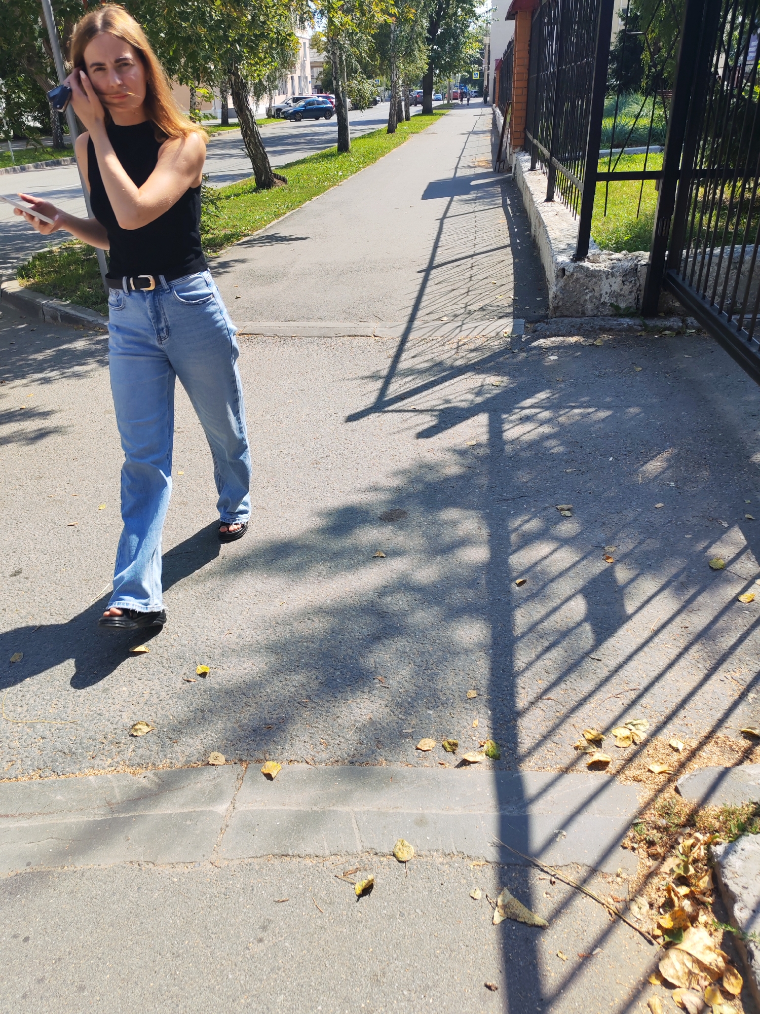 A blonde woman wearing a black sleeveless top, light blue flared jeans, a black belt with a gold buckle, and black platform sandals walks on a paved sidewalk. She is holding a smartphone in her left hand and appears to be adjusting her hair with her right hand. The sidewalk is adjacent to a street with cars in the background and a grassy area with trees to her left. To her right, there is a black metal fence. The ground is partially covered with fallen leaves, and the scene is illuminated by sunlight casting strong shadows on the pavement.