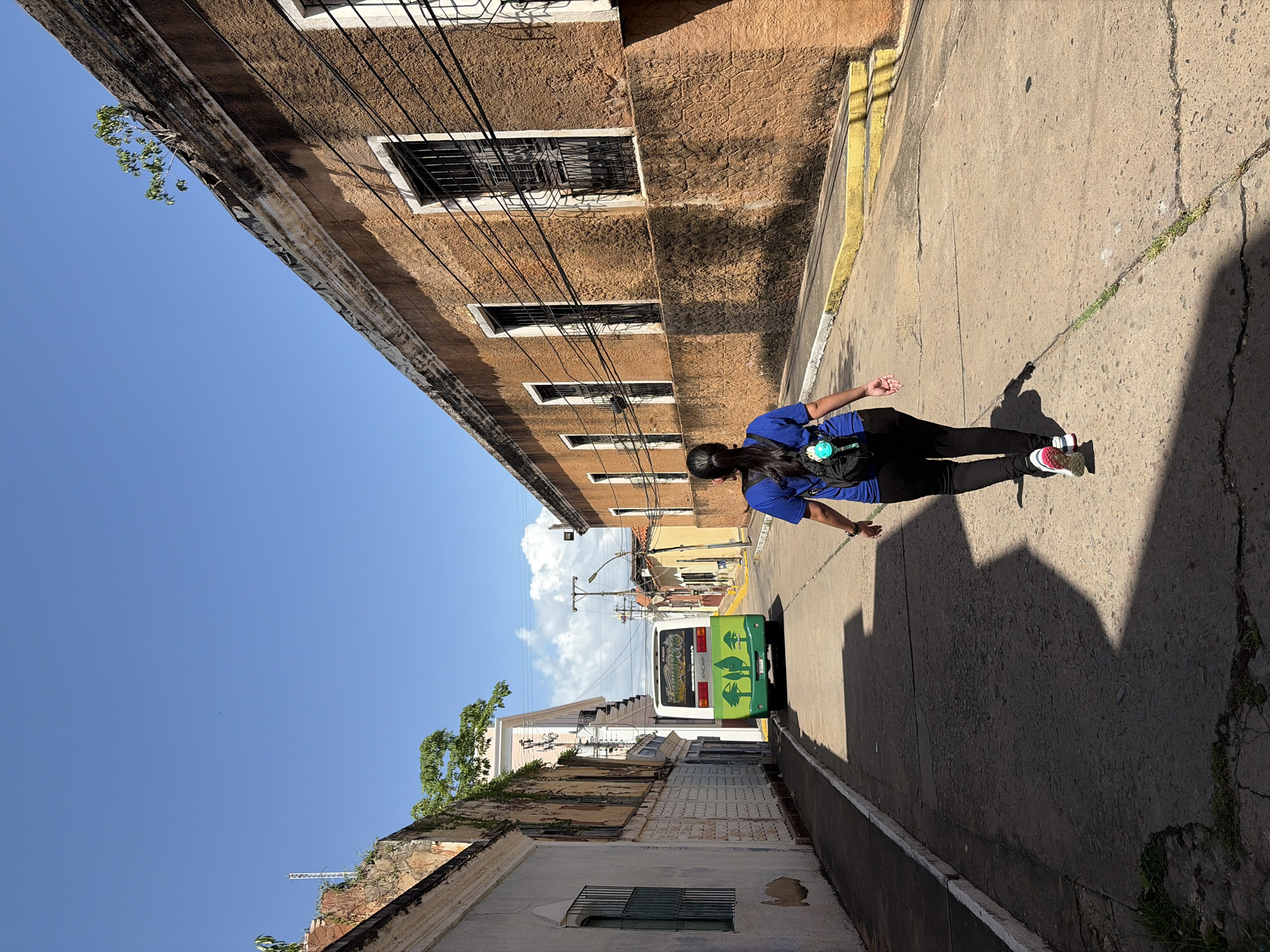 The image shows an urban street scene in Ciudad Bolivar, Venezuela. A person wearing a blue shirt and black pants walks along the sidewalk. Their shadow is visible on the concrete. Buildings line both sides of the street, featuring a mix of architectural styles and conditions. Some buildings have windows with bars, while others show signs of age and decay. Electrical wires crisscross the sky above the street. In the distance, a green and white bus is visible. The sky is clear and blue.