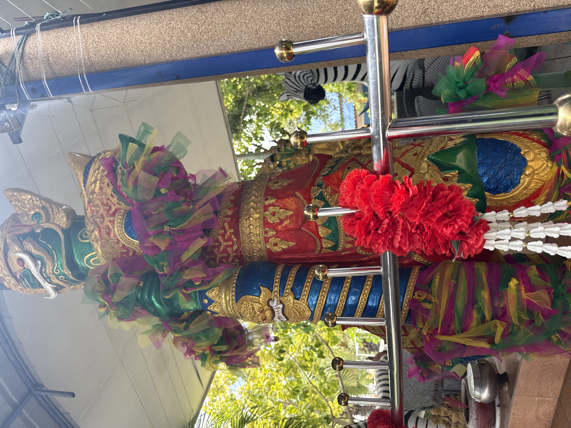 An outdoor scene in Bang Sare, Thailand, features a vibrantly decorated, large green and gold statue of a mythical guardian figure. The statue is adorned with colorful fabric streamers in shades of pink, yellow, and green, and a red flower garland hangs in the foreground. In the background, a black and white zebra statue is visible, along with lush green trees and foliage under a covered structure with a corrugated roof. The scene is likely part of a temple or cultural site.