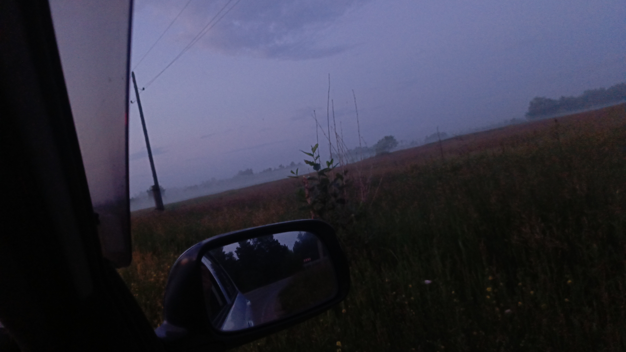 The image depicts a rural landscape at dusk, viewed from inside a vehicle. A side mirror reflects a road lined with trees. The sky is a muted purple and blue, with hints of clouds. A power line pole stands prominently in the background, suggesting the presence of infrastructure in the area. The terrain appears to be agricultural, with fields of grass and some scattered vegetation. The overall atmosphere is serene and slightly melancholic, possibly indicative of early morning or late evening hours. The image does not provide any specific visual cues to pinpoint its location within Angarsk, Russia.