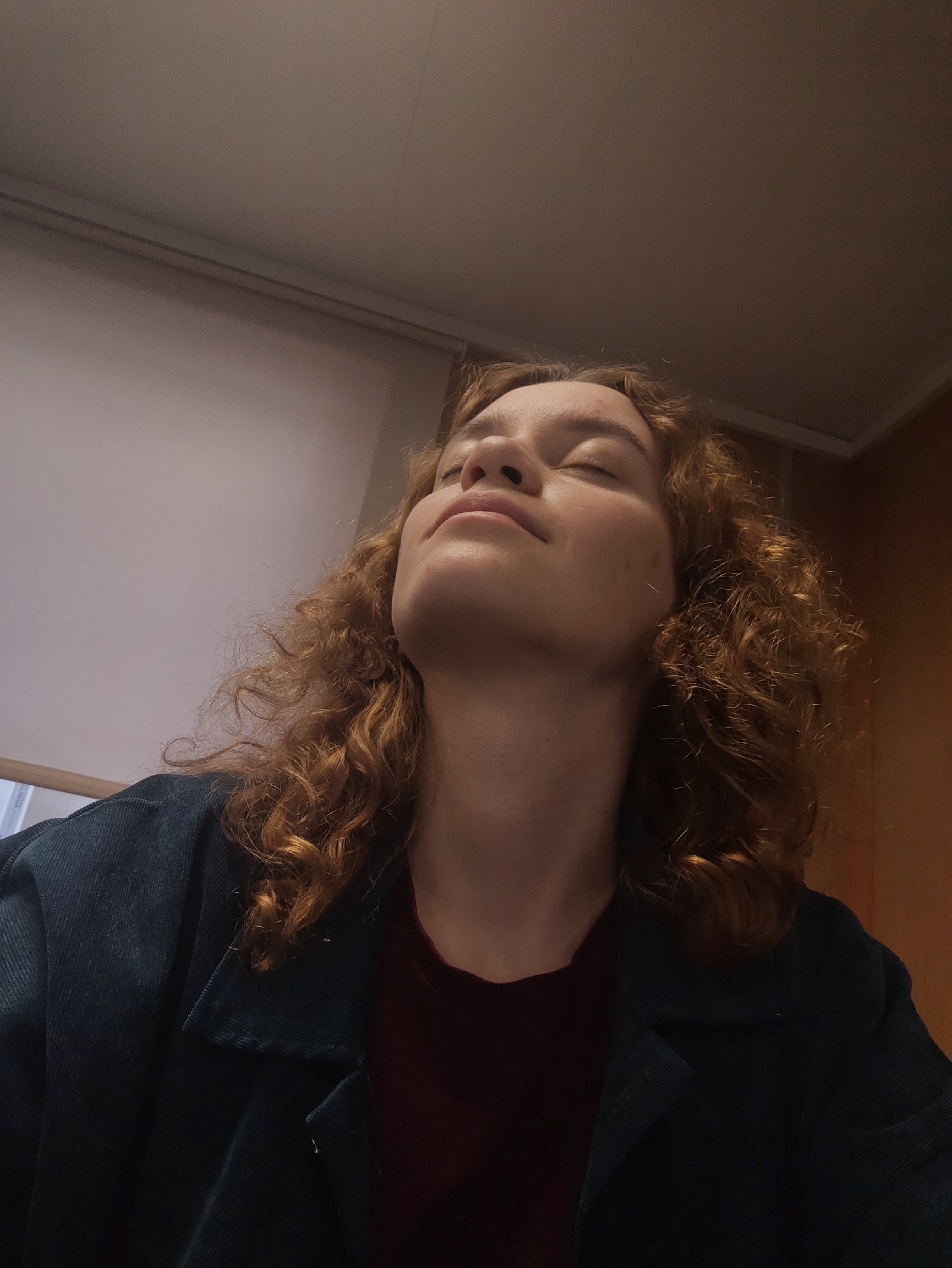 A woman with curly, reddish-brown hair is looking upwards with her eyes closed. She is wearing a dark teal corduroy jacket over a maroon shirt. The background includes a white roller blind and a light brown wall. The location is Unknown, Unknown.