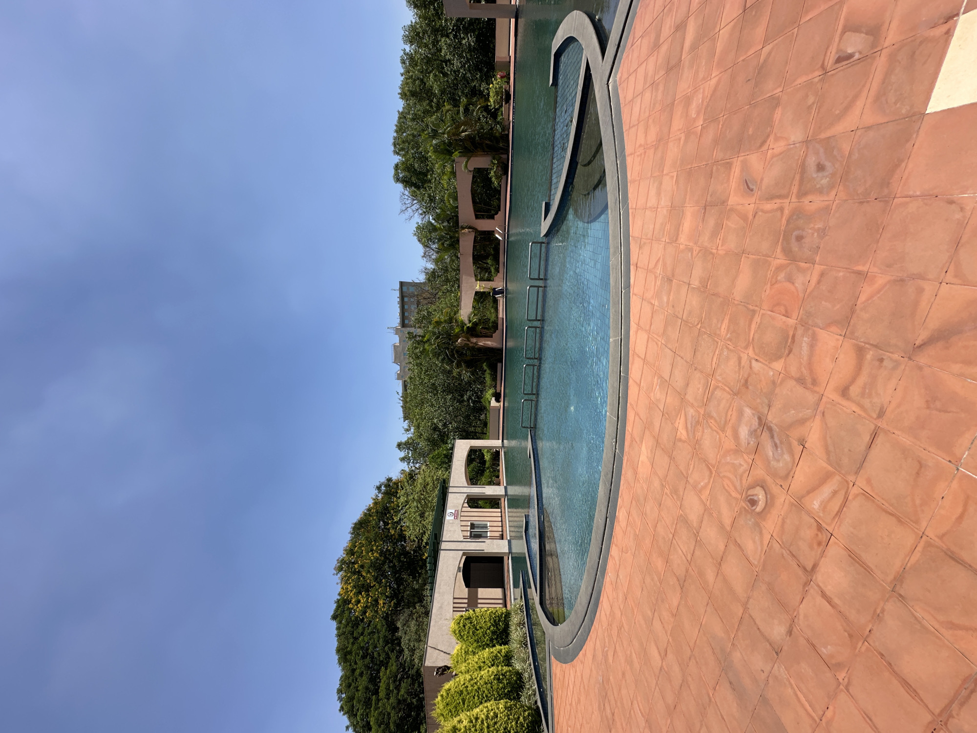The image shows an outdoor swimming pool at an angle. The pool's edge is bordered by reddish-brown tiles. The pool itself is a complex shape with varying depths indicated by the color of the water. Metal ladders are visible within the pool. Beyond the pool are trees and architectural structures with arched openings. The sky is blue. The location is Marunji, India.
