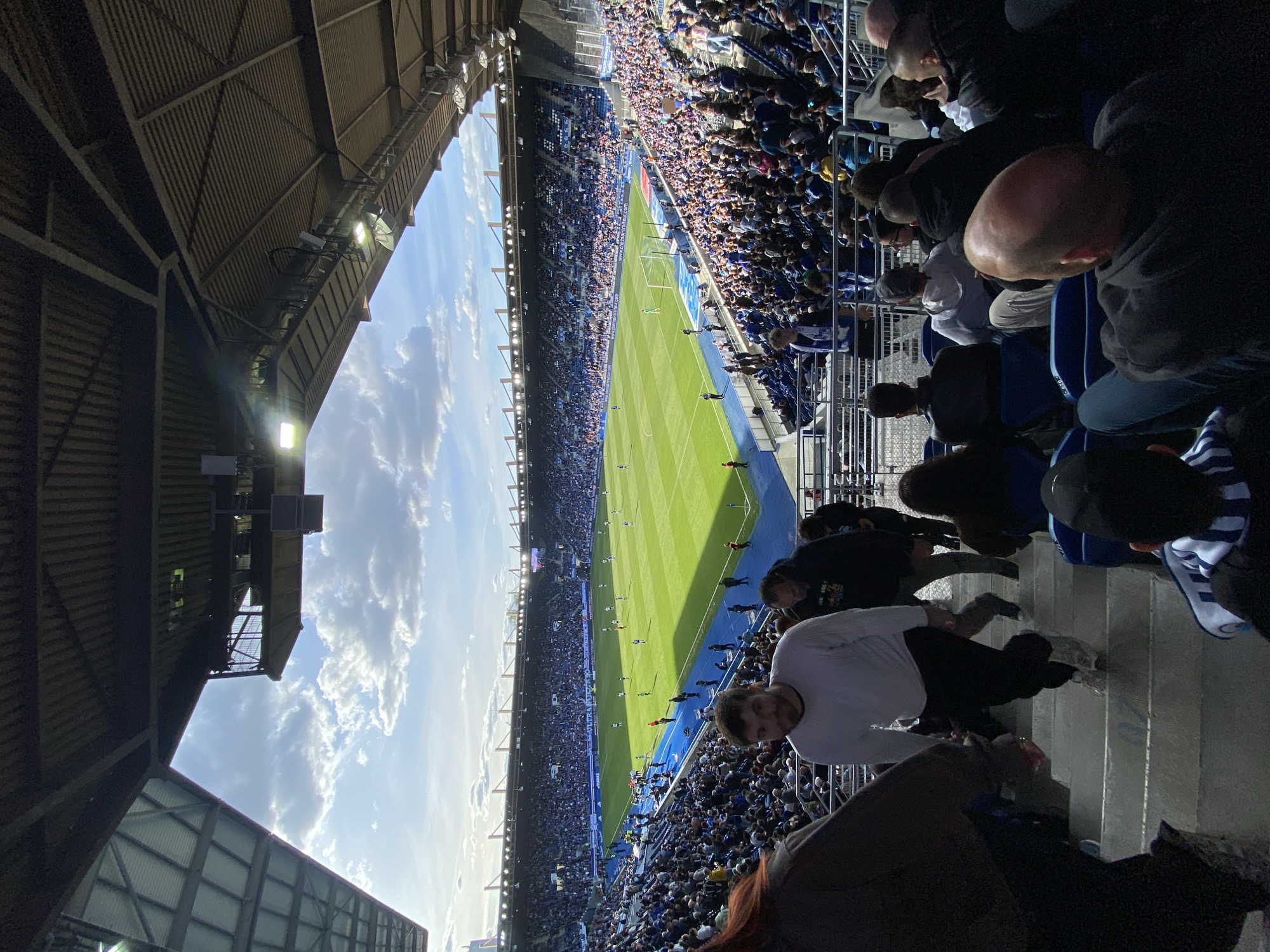 The image shows a view from the stands of a soccer stadium, likely the Stade de la Meinau in Strasbourg, France, given the blue-and-white colors associated with RC Strasbourg Alsace. The field is visible, filled with players and the game in progress. The stands are densely packed with spectators wearing a mix of clothing, predominantly in blue. People are seated or standing in the rows of the stands, facing towards the field. Overhead, the stadium roof structure is visible. The sky above the stadium is blue with some clouds. A person in a white shirt looks towards the camera, walking up stairs.