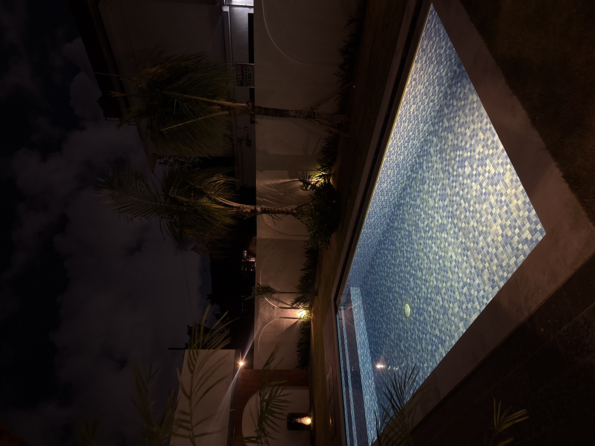 This nighttime image depicts an outdoor area, likely a resort or villa garden, in North Kuta, Indonesia. The scene is dominated by a brightly lit swimming pool on the right side, tiled with a mosaic pattern of blue and white squares. The water reflects the artificial lights, creating a luminous effect within the pool.
To the left of the pool, several tall palm trees with visible trunks and fronds rise against a dark, cloudy night sky. The trees and surrounding foliage are illuminated by strategically placed ground-level spotlights, casting warm light on the lower parts of their trunks and the lush greenery. Behind the palm trees, modern, light-colored architectural walls with distinct curved indentations are visible, also highlighted by the artificial lighting. A building structure is partially visible further in the background on the far left.
The overall impression is one of a serene and well-maintained tropical environment, experienced after dusk, with artificial illumination creating a dramatic contrast between the glowing pool and garden elements against the deep darkness of the night sky. No people or visible text are present in the image.