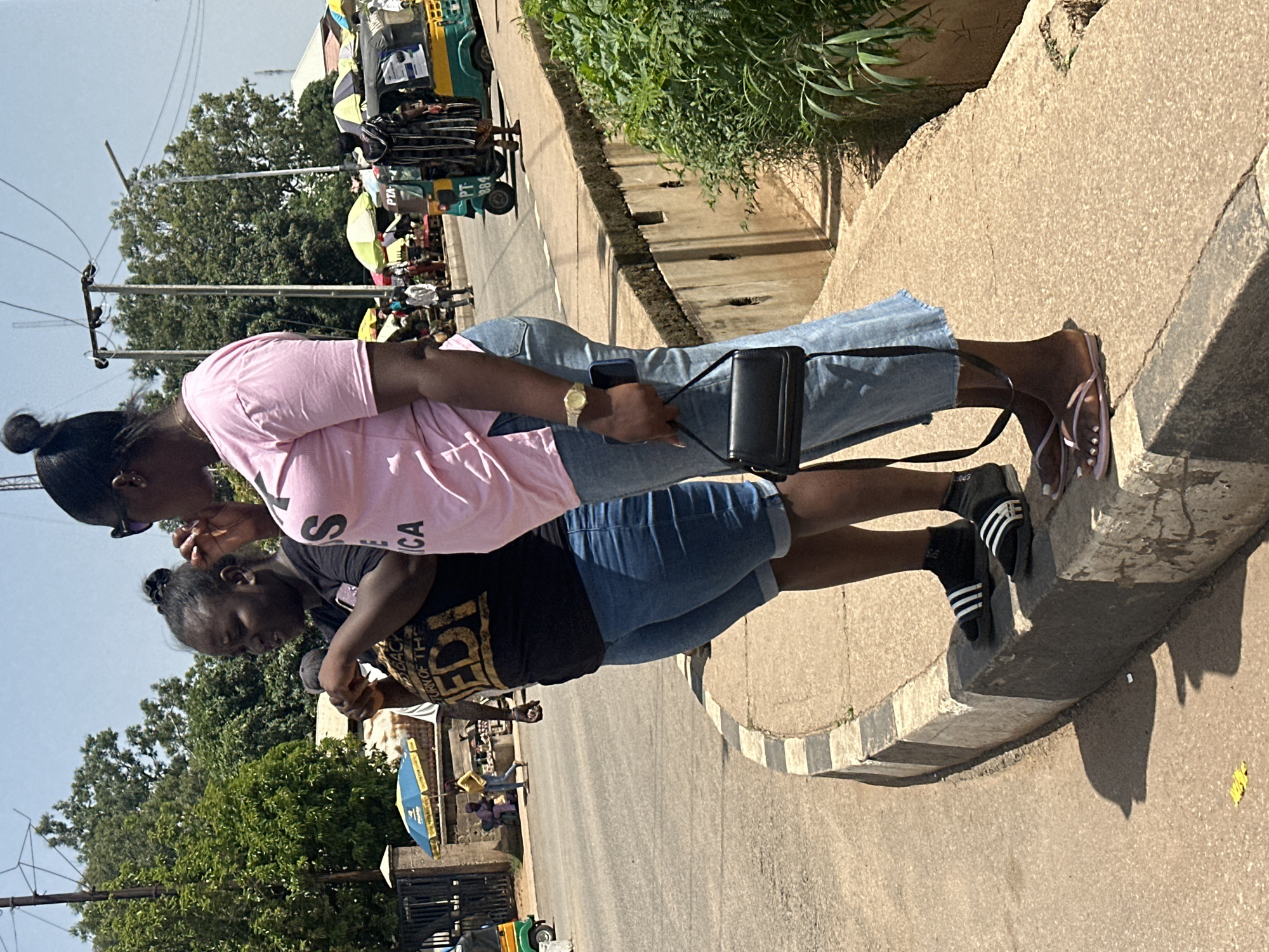 Two women are standing on the side of a road in Barakin Lerawi, Nigeria, in what appears to be a marketplace. The woman on the left wears a black t-shirt with a graphic print and blue denim shorts. She is holding something in her hands and looking down. The woman on the right wears a pink t-shirt and light blue denim jeans. She has sunglasses on her head, is holding a black handbag, and appears to be talking or eating. In the background, there are various stalls with umbrellas, vehicles including an auto-rickshaw, and trees. The ground is paved with asphalt and a concrete curb.