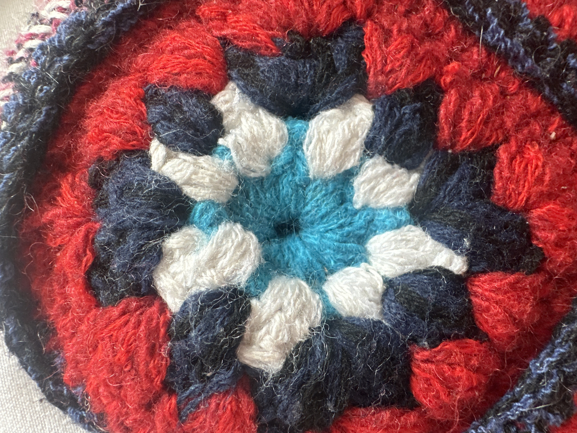 This media file is a close-up, overhead shot of a crocheted circular object with a star-like pattern in the center. The object is made from yarn in various colors, including bright red, navy blue, white, and a light turquoise or cyan. The texture of the yarn is fuzzy, with some strands appearing looser than others. The crocheting technique creates distinct stitches and raised areas, forming a geometric design. The pattern radiates outwards from a central point, with alternating colors and shapes.
The outer edge of the crocheted object features a border in navy blue and black, which appears to be a different crocheting style, possibly a denser stitch or multiple strands. The background is a light, textured surface, likely a table or fabric.
No people, activities, time of day, weather, or location cues are visible. There is no visible text in the image. The object appears to be a handmade craft, possibly a coaster, a decorative element for clothing, or a component of a larger textile project. The colors are vibrant and the pattern is intricate, suggesting careful craftsmanship. The overall impression is one of texture and color.