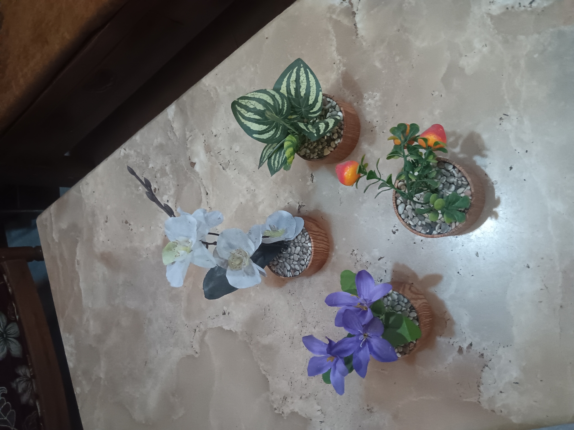 Four artificial potted plants are arranged on a marble-patterned surface. The plants include a white orchid, a purple flowering plant, a striped green plant, and a plant with orange and red fruit-like decorations. The marble surface appears to be a tabletop. In the background, a dark brown wooden chair is visible. The scene is indoors.