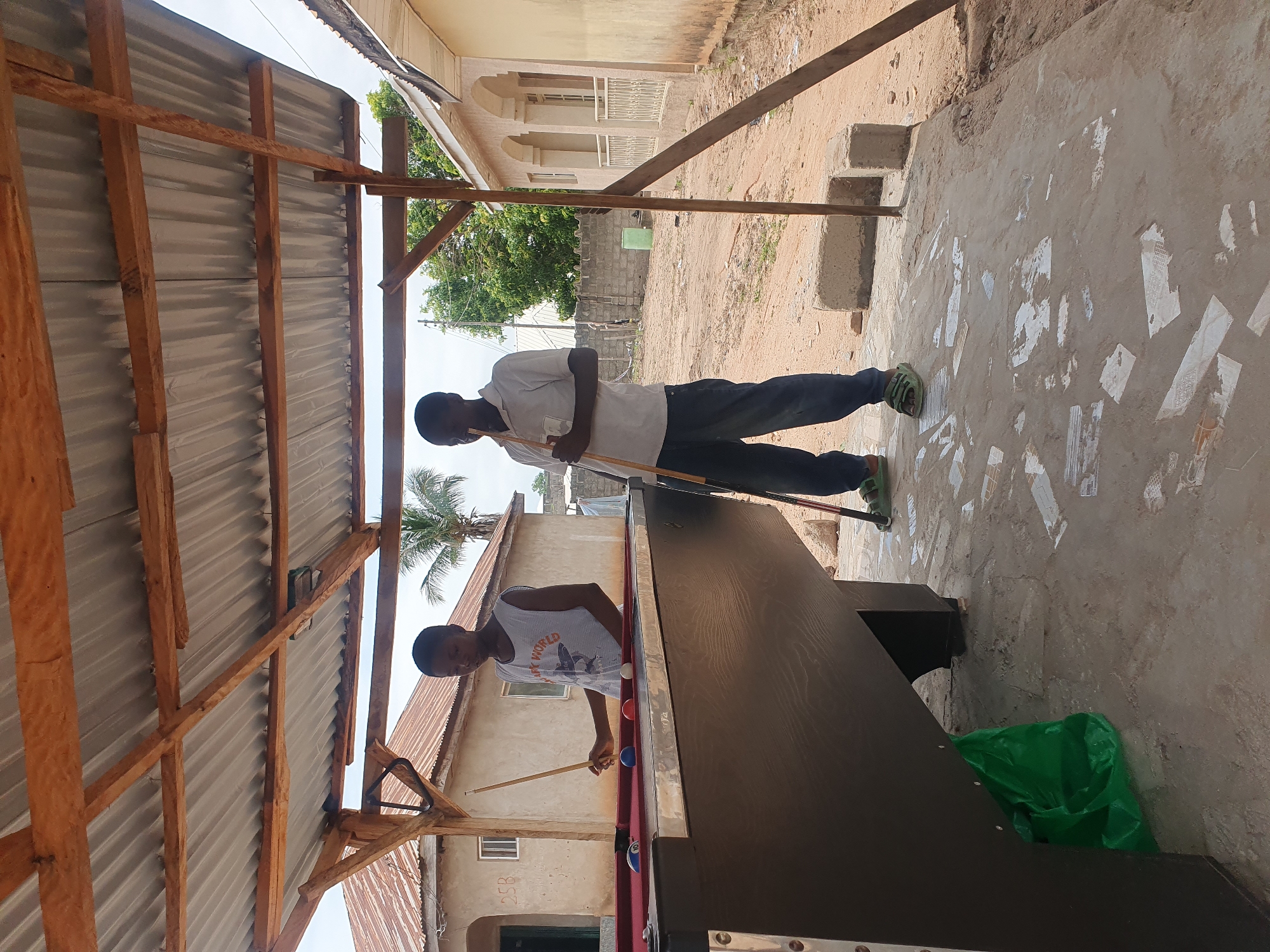 In Lokoja, Nigeria, two young boys are playing pool under a makeshift shelter with a corrugated metal roof supported by wooden beams. The shelter is constructed over a concrete pad. A dark-colored pool table with a red felt surface is situated on the pad. Several billiard balls, including a blue ball with the number 10, a solid blue ball, a red ball, and a white cue ball, are visible on the table. The boy on the left, wearing a grey sleeveless shirt with a shark graphic, holds a pool cue. The boy on the right, dressed in a white polo shirt and dark trousers, also holds a pool cue. In the background, a modest building with a corrugated metal roof and simple windows is visible, along with a palm tree. The ground around the pool table is sandy and uneven, with scattered pieces of broken tiles. A bright green plastic bag is placed underneath the pool table. The overall scene depicts an outdoor recreational area in a residential setting.