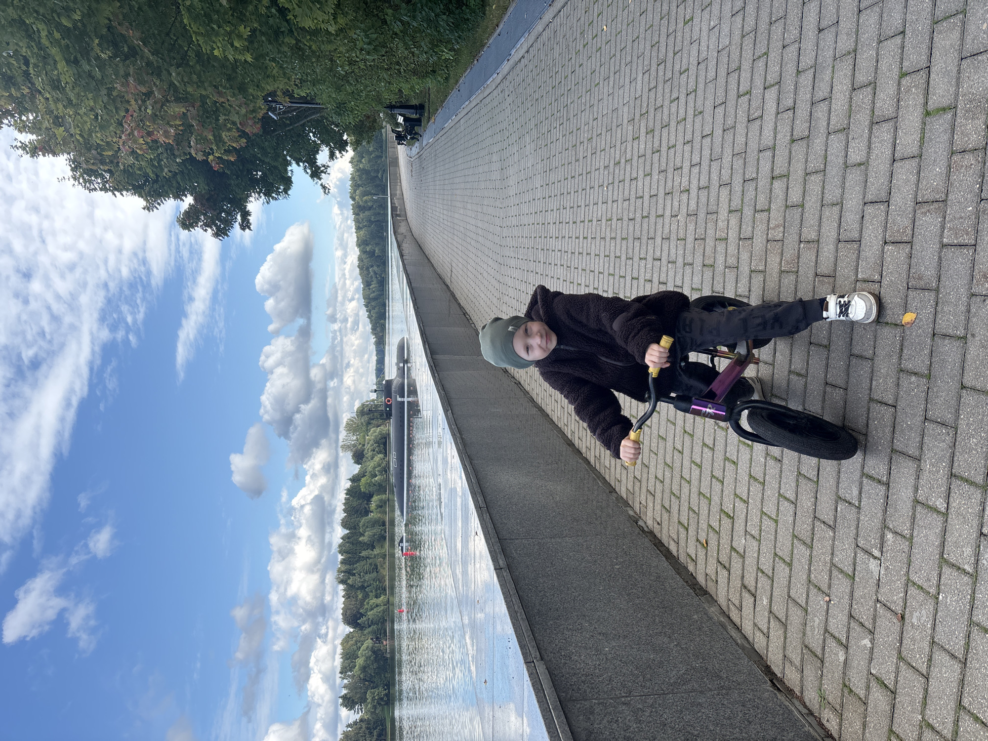 The image shows a young person on a balance bike on a paved pedestrian walkway next to a body of water. In the background, a submarine is visible in the water, and a line of trees forms the distant shoreline. The walkway is made of rectangular grey pavers, and there is a granite wall separating the walkway from the water. The sky is partly cloudy with a blue sky. The location appears to be the embankment in Рождествено, Russia, a known location for a naval base.