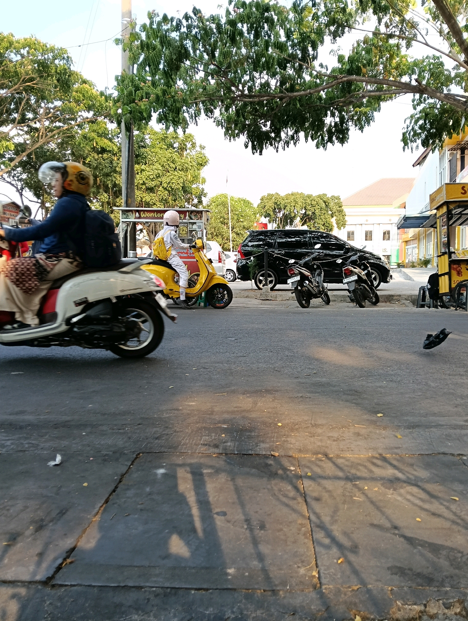The image depicts a street scene in Karanganyar, Indonesia, characterized by the presence of several motorcycles and a black SUV. In the foreground, a white scooter carries two individuals; the rider wears a dark jacket and backpack, and the passenger is obscured by the rider. To the right, a yellow scooter is ridden by a person in white attire and a pink helmet, carrying a yellow backpack. The scooters are moving on a paved road, with shadows of trees and possibly a fence falling across the surface.
In the background, a black SUV is parked on the side of the road. Adjacent to it, there are two smaller motorcycles. Further to the right, a yellow food stall with signage that reads "Wonton" is visible, along with some chairs and tables. Buildings with white facades and tiled roofs line the street. Lush green trees and foliage are abundant, suggesting a somewhat tropical environment. The sky is bright and appears to be daytime. The overall impression is a typical urban street in Indonesia with everyday transportation and commercial activity.