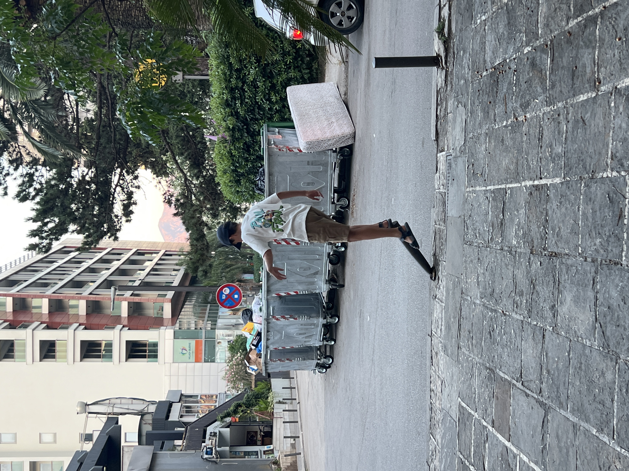 The image depicts a street scene in Budva, Montenegro. In the foreground, a person wearing a cap, t-shirt, and shorts is seen standing on a street, with their left foot on a black object, possibly a skateboard or scooter, in a precarious balancing act. Behind this individual are several large, overflowing trash dumpsters. A white mattress is leaning against one of the dumpsters. In the background, a multi-story building with a modern architectural design stands, featuring balconies and windows. Trees and foliage are also visible, suggesting an urban environment with green spaces. The lighting indicates it is daytime, with a soft glow suggesting it could be late afternoon or early morning.